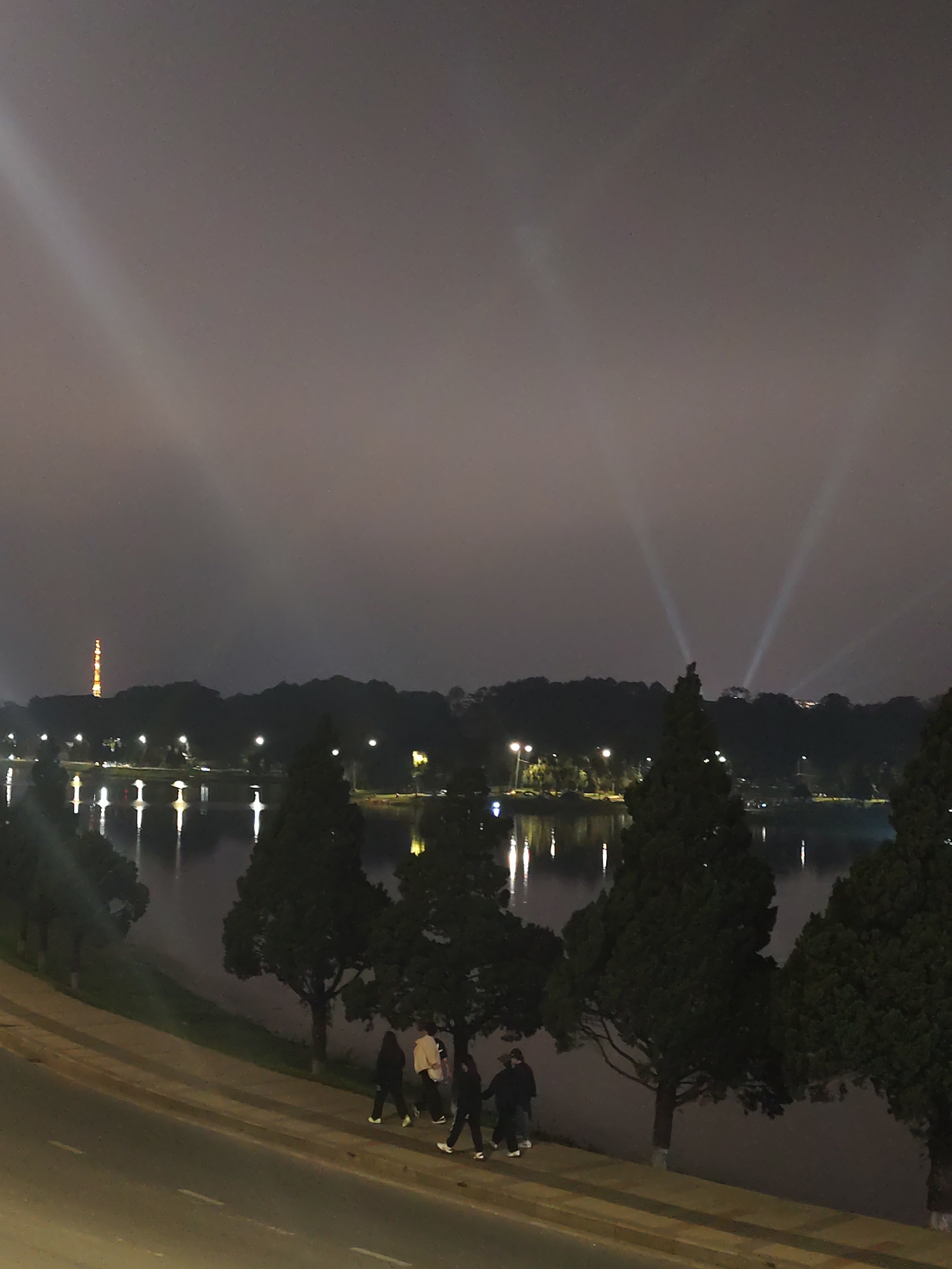 The image displays a nighttime scene in Da Lat, Vietnam, likely near Lâm Viên. In the foreground, a group of at least four individuals is walking along a sidewalk that runs parallel to a road. Tall, dark conifer trees line the edge of the sidewalk, separating it from a calm body of water, presumably a lake, which occupies the middle ground.
Artificial lights from the far shore of the lake are brightly reflected on the water's surface, creating distinct vertical streaks of illumination. The distant shoreline is also dotted with numerous small lights. In the background, rolling hills rise under a dark, overcast or misty sky. Prominently featured are several powerful spotlights projecting bright, conical beams of light upwards into the sky. On the far left of the horizon, a distinct orange-lit tower, characteristic of the Da Lat TV tower, stands tall against the dark sky. The overall atmosphere is peaceful and illuminated by the various artificial lights, indicative of an urban or park setting at night. There is no visible text in the image.