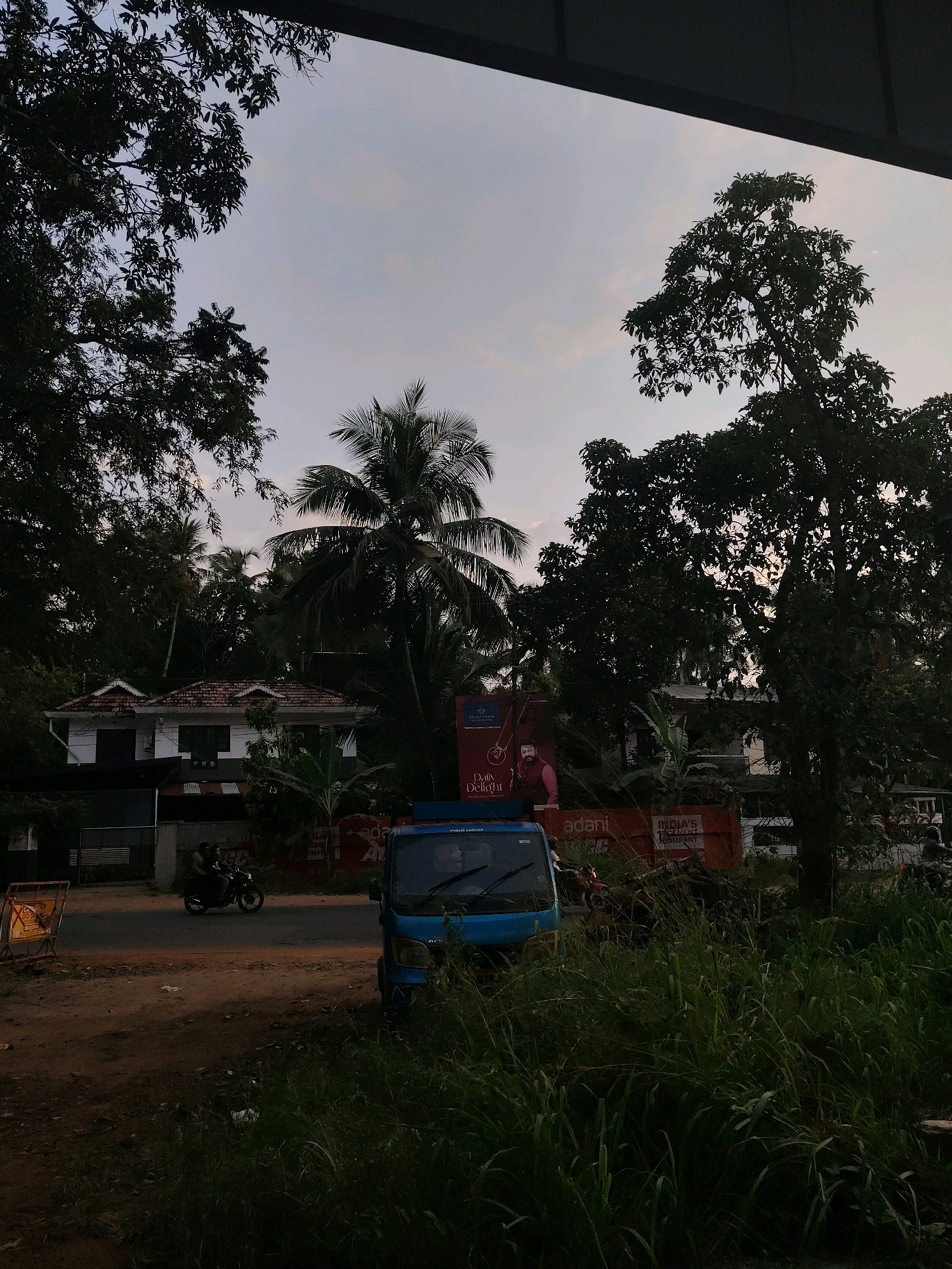 The image depicts a street scene in Makkaraparambu, India, during daylight. A blue truck is parked on the right side of the frame, partially obscured by tall green grass. To the left of the truck, a road runs horizontally. On the road, two individuals are riding a motorcycle in motion, heading towards the left. Further left, a white building with a tiled roof stands behind a dark fence. Dense foliage and trees surround the scene, with a coconut palm visible in the center. A large billboard with images and text is situated behind the truck and to the right of the building. The sky above is overcast with diffused light, suggesting either dawn or dusk. The overall impression is that of a quiet, possibly rural or semi-urban, environment with everyday activity.