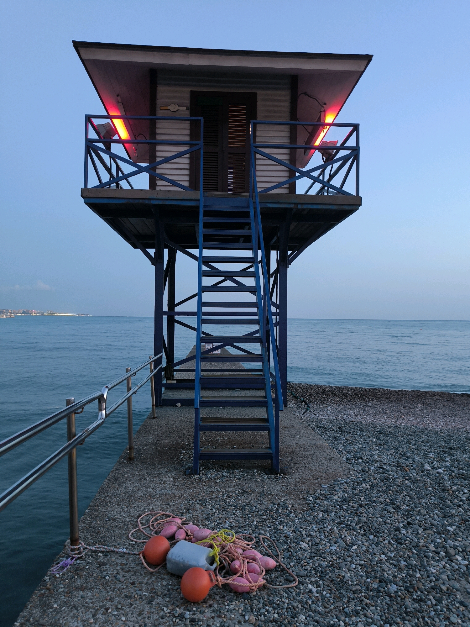 A tall, blue metal structure supporting a small, white lifeguard station with a sloped roof is situated on a pebble beach. Two red lights illuminate the underside of the roof. A blue metal staircase leads up to the station, which has a closed door and railing. In the foreground, a coiled rope with pink and orange buoys lies on the concrete and pebble surface. The ocean stretches out to the horizon, with a faint city skyline visible in the distance on the left. The sky is a pale blue, indicating either dawn or dusk. The scene is located on the coast of Sochi, Russia.