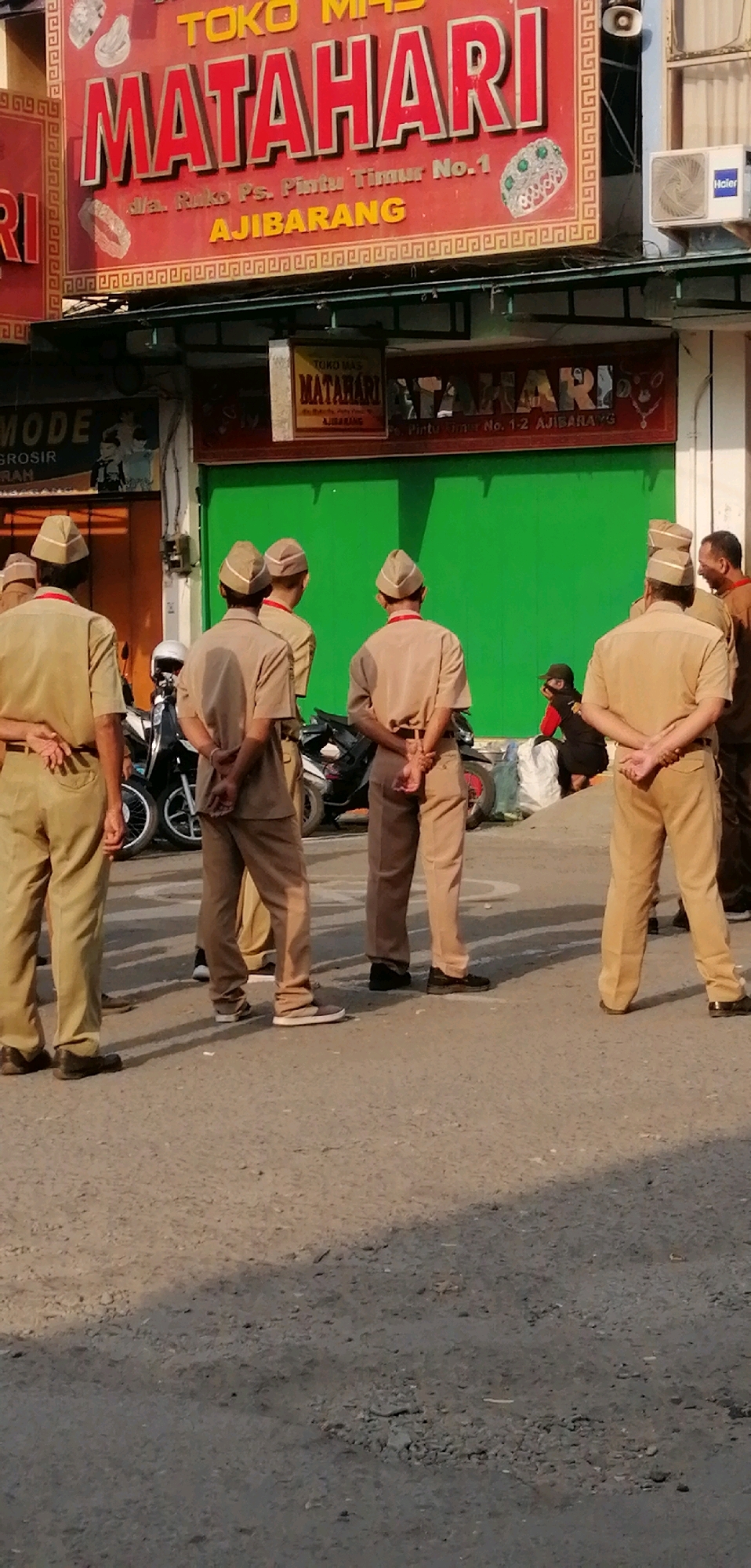 The image captures a street scene in Ajibarang, Indonesia, featuring several individuals in uniform standing in front of a building with a prominent red and gold sign that reads "TOKO MAS MATAHARI AJIBARANG". The sign also displays an address: "Jl. Raya Ps. Pintu Timur No.1". The individuals appear to be male, wearing light brown uniforms with matching hats. Some have their hands clasped behind their backs, while others are standing with their arms at their sides. In the background, motorcycles are parked, and a bright green wall forms a backdrop. There are also other signs visible, including one that says "MODE GROSIR" and another smaller "MATAHARI" sign. The overall environment suggests a commercial area.
