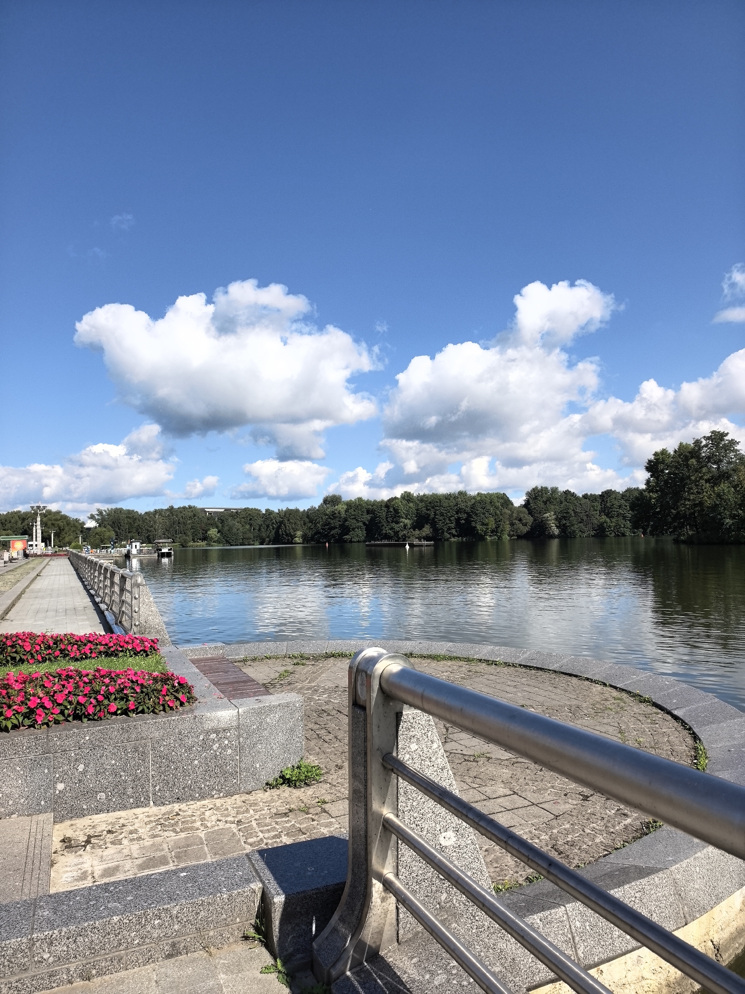 A long shot captures a sunlit, serene waterfront park scene under a vast, blue sky dotted with puffy white clouds. The foreground features a paved walkway with a polished metal railing. To the left, a planter box overflows with vibrant pink flowers, set against a textured granite wall. Beyond this, the walkway extends towards a distant line of lush green trees that form the far bank of a calm body of water. Several small boats are visible moored near the shore. The water reflects the blue sky and white clouds, creating a peaceful, bucolic atmosphere. The scene appears to be in an urban park, with hints of lampposts and structures visible along the walkway in the distance, suggesting a developed public space.
