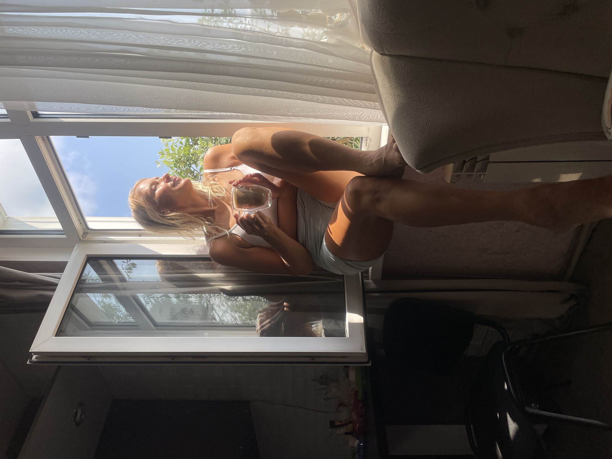 A blonde woman sits on a windowsill, holding a glass mug of coffee. She is wearing a white bra and light-colored shorts. The window is open, revealing greenery and a blue sky outside. Sunlight streams into the room, casting shadows. To the left of the window, a dark chair and a shelf with various items are visible. To the right, sheer white curtains hang, and a textured armchair is partially visible. The scene appears to be an indoor setting in a residential area. There are no discernible features in the image that would allow for identification of the specific location within Baranovka, Russia.