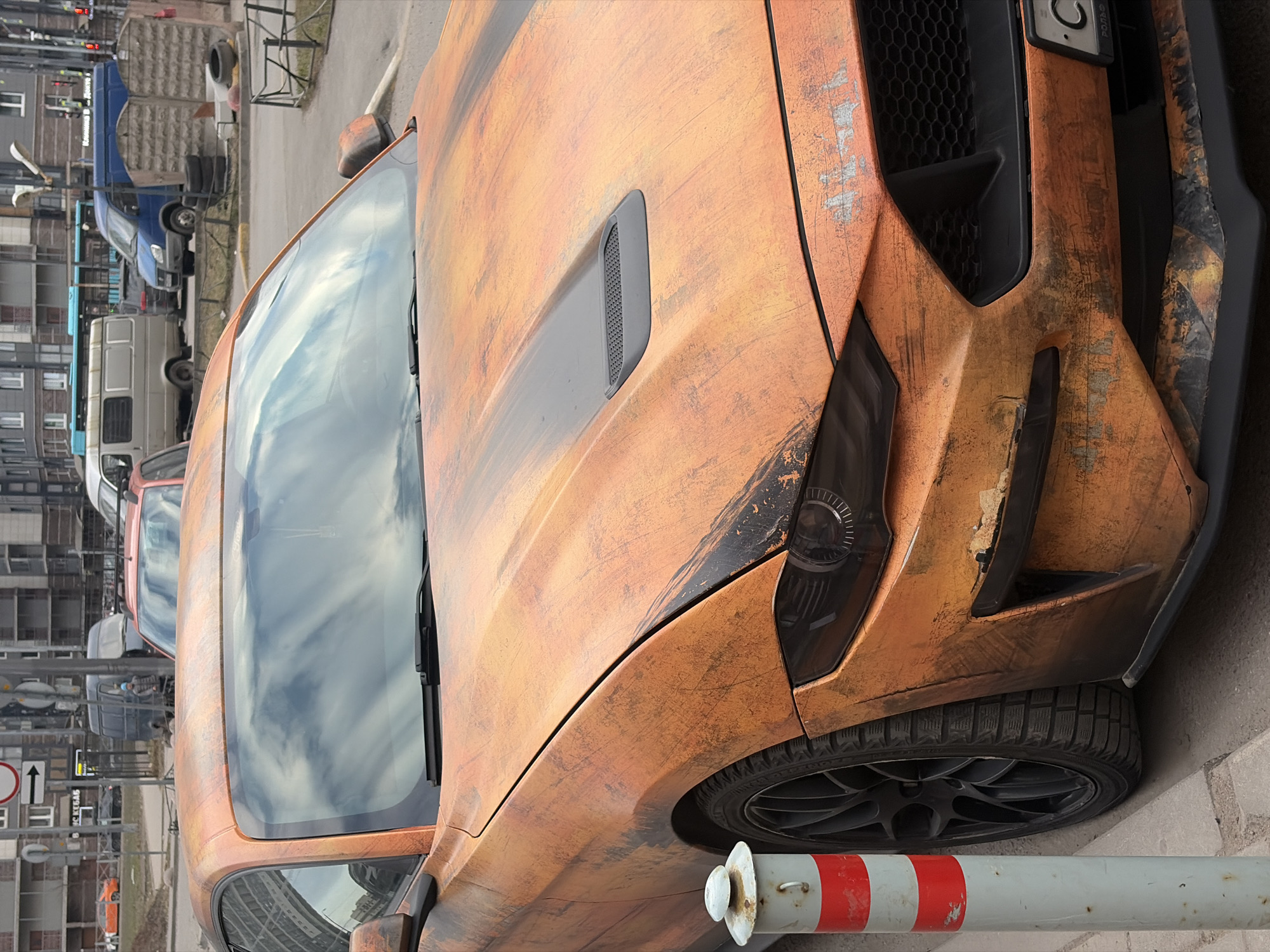 The image shows a close-up of an orange, textured car, possibly a Ford Mustang, parked on a street. The car has a black grille and black wheels. The license plate is partially visible. The street scene in the background includes various vehicles, such as vans and cars, parked along the road. There is a building with a gray exterior in the background. Street signs and traffic control devices are visible. A red and white striped pole is in the foreground near the car's tire. Based on the street signs and vehicle types, the location is identified as Saint Petersburg, Russia.