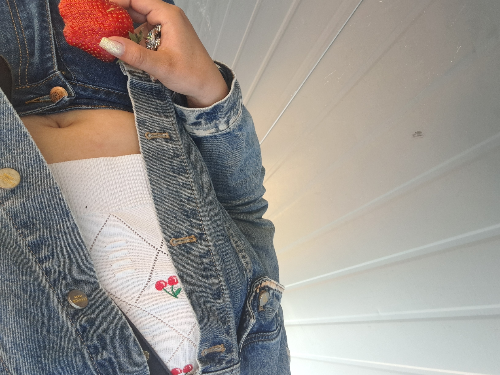 A person wearing a denim jacket and a white top with cherry embroidery is holding a strawberry in their hand. The person is standing in front of a white, grooved wall.