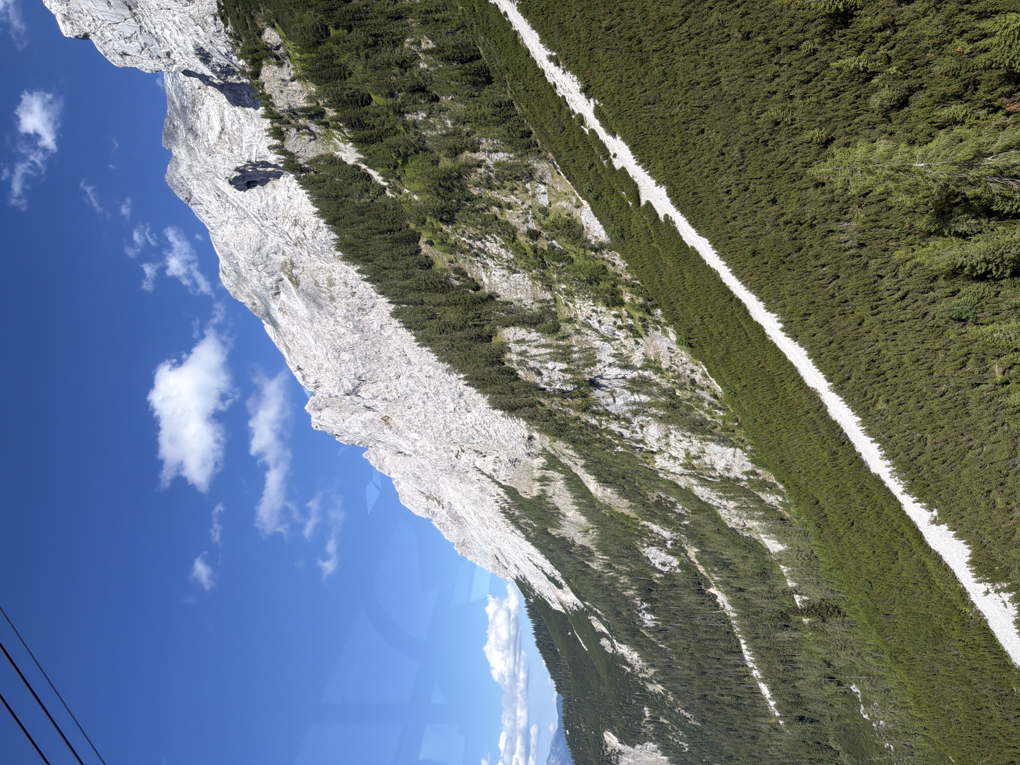 The image depicts a mountainous landscape under a clear blue sky with scattered white clouds. In the foreground, a dense forest of green pine trees covers a steep slope, with a narrow, light-colored path winding its way through them. The path appears to be made of gravel or small stones. Further up the slope, the forest gives way to rocky, lighter-colored mountainside. The sheer rock face of the mountain dominates the upper portion of the image, showing intricate geological formations and textures. The scene conveys a sense of natural beauty and rugged terrain, likely in an alpine region. It is not possible to determine the specific location within Grainau, Germany from the visual information provided.