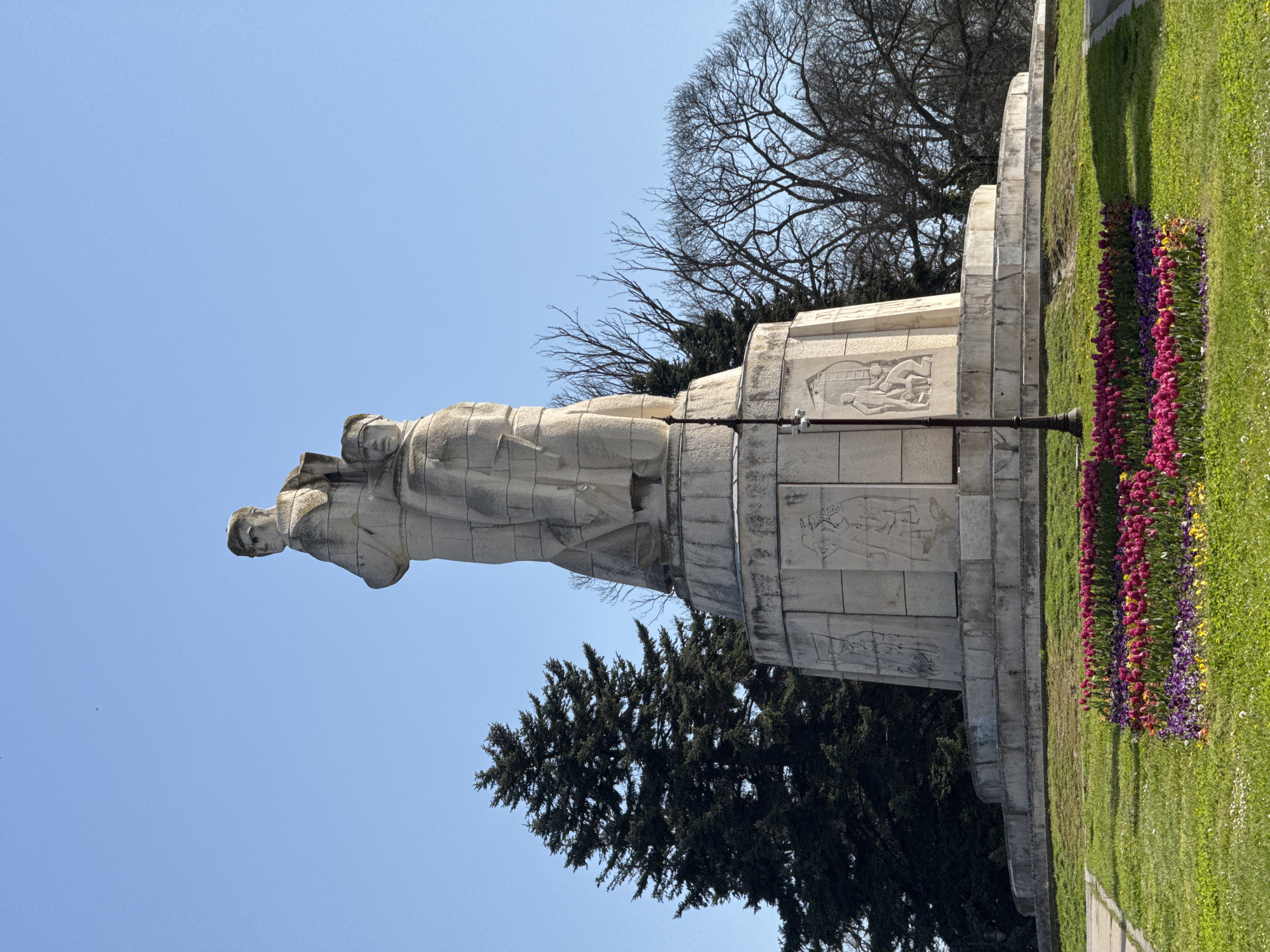 The image displays a tall, light-colored stone monument standing prominently against a clear, vibrant blue sky in what appears to be a well-maintained park or memorial area in Varna, Bulgaria. The central feature of the monument is a group of figures, likely soldiers or commemorative figures, carved in a heroic or solemn posture, standing atop a large, multi-tiered pedestal.
The pedestal itself is ornate, featuring bas-relief carvings on its sides that depict various scenes with human figures, though their specific narratives are not distinctly clear. A dark, slender lamp post is affixed to the lower section of the monument's base.
The ground around the monument is covered in lush green grass, and vibrant flower beds are arranged in neat rows along the base of the monument. These beds display a rich array of colors, with distinct sections of red, pink, yellow, and purple flowers. In the background, a mix of trees can be seen: some with their dark, bare branches silhouetted against the bright sky, suggesting a deciduous type in early spring or late autumn, and others with dense, dark green foliage, indicative of evergreen species. The scene is brightly lit by the sun, indicating daytime. No visible text can be identified on the monument or elsewhere in the image. The setting evokes a sense of solemnity and public remembrance.