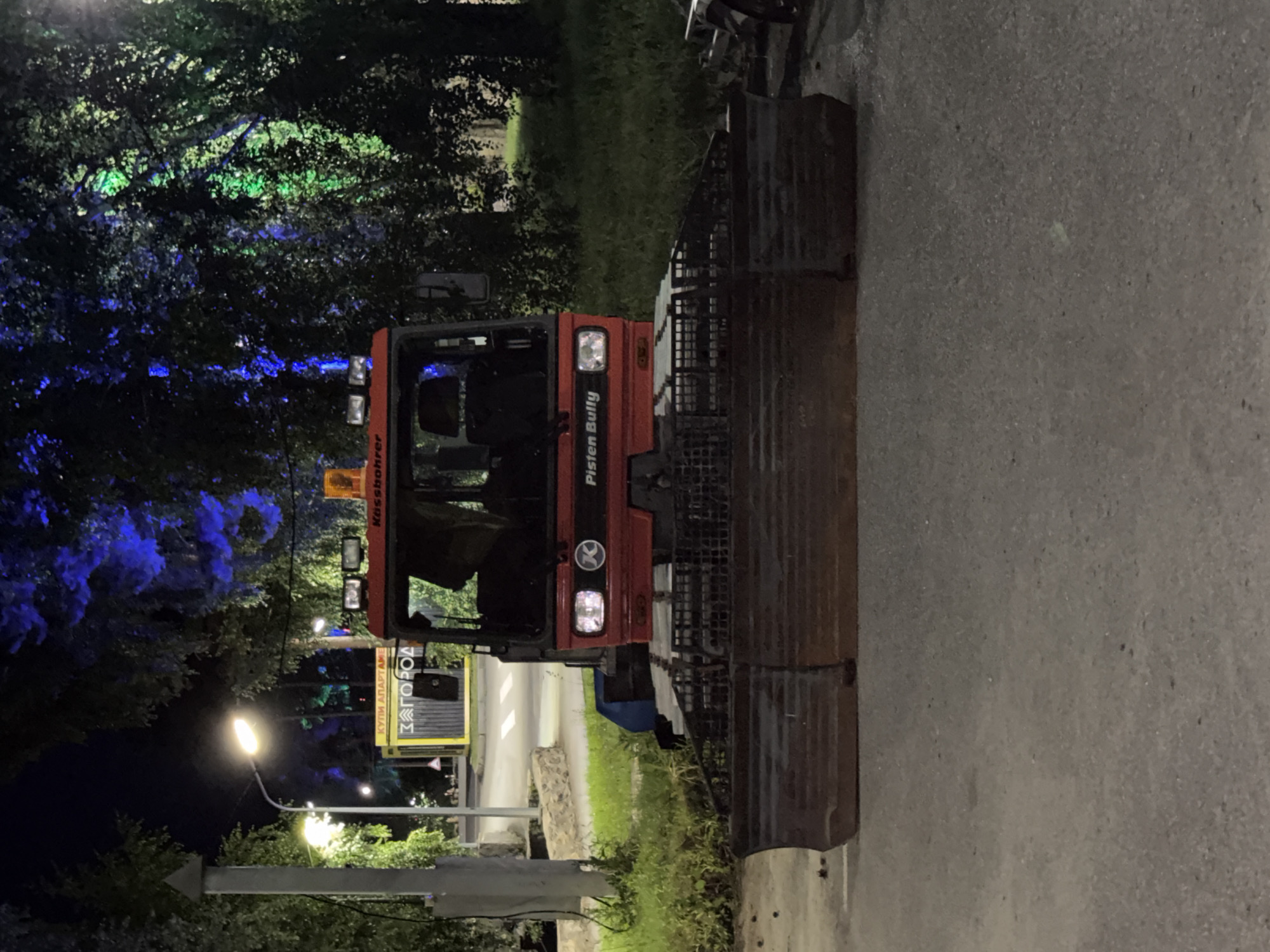 A Pisten Bully vehicle is parked on a paved surface at night, with trees and lighting in the background. The vehicle is red and white, with a large metal plow attachment in the front. The cab of the vehicle is visible, with seats inside. In the background, trees are illuminated with blue and green lights. A yellow kiosk with Russian text "КУПИ АПАРТАМЕНТЫ" (Buy Apartments) and "ЗАГОРОД" (Suburb) is visible to the left of the vehicle. The image appears to be taken in the city of Turgoyak, Russia, based on the kiosk text and the general appearance of the scene. There are no people visible in the image.