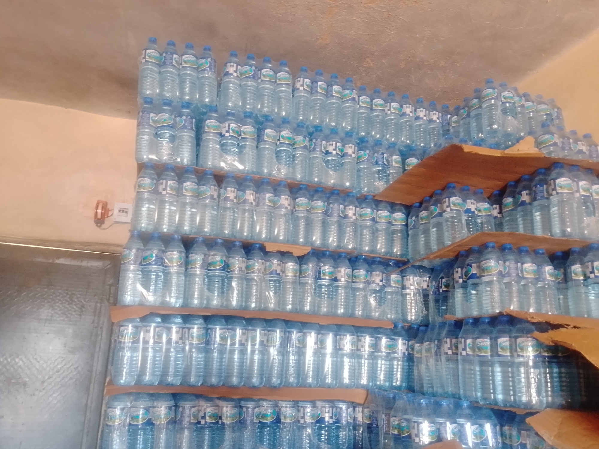 The image displays a large, organized stockpile of bottled water. The bottles are clear plastic with blue caps and feature white labels with green and blue accents. The water inside appears clear. The bottles are stacked neatly on wooden shelves, creating multiple rows and columns that fill the frame. The lighting suggests an indoor setting, possibly a warehouse or storage area, with a plain, textured ceiling above. The overall impression is one of commercial inventory or preparation for distribution. There are no people visible in the image. The activity taking place is the storage of goods. Notable details include the sheer volume of identical water bottles, suggesting a commercial purpose. The setting appears to be a functional storage space with basic shelving. No specific time of day or weather conditions can be determined from the image. No text is legible on the bottle labels due to the resolution.
