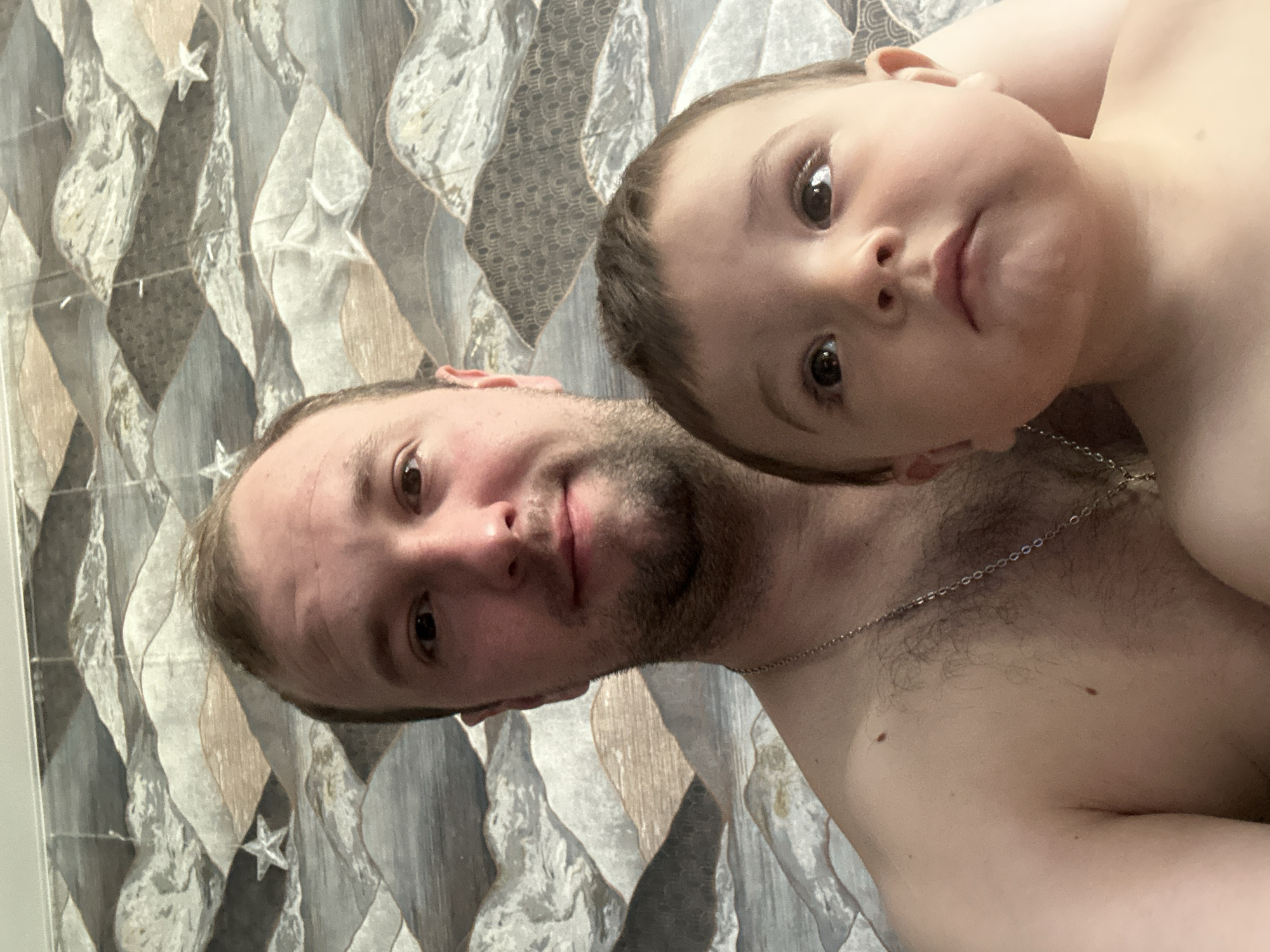 A shirtless man with fair skin and a beard lies next to a young child, both facing the camera. The child is fair-skinned and appears to be resting its head on the man's chest. The man wears a thin metal chain around his neck. The background appears to be a wall with a patterned covering. The location is Chaikovsky, Russia.