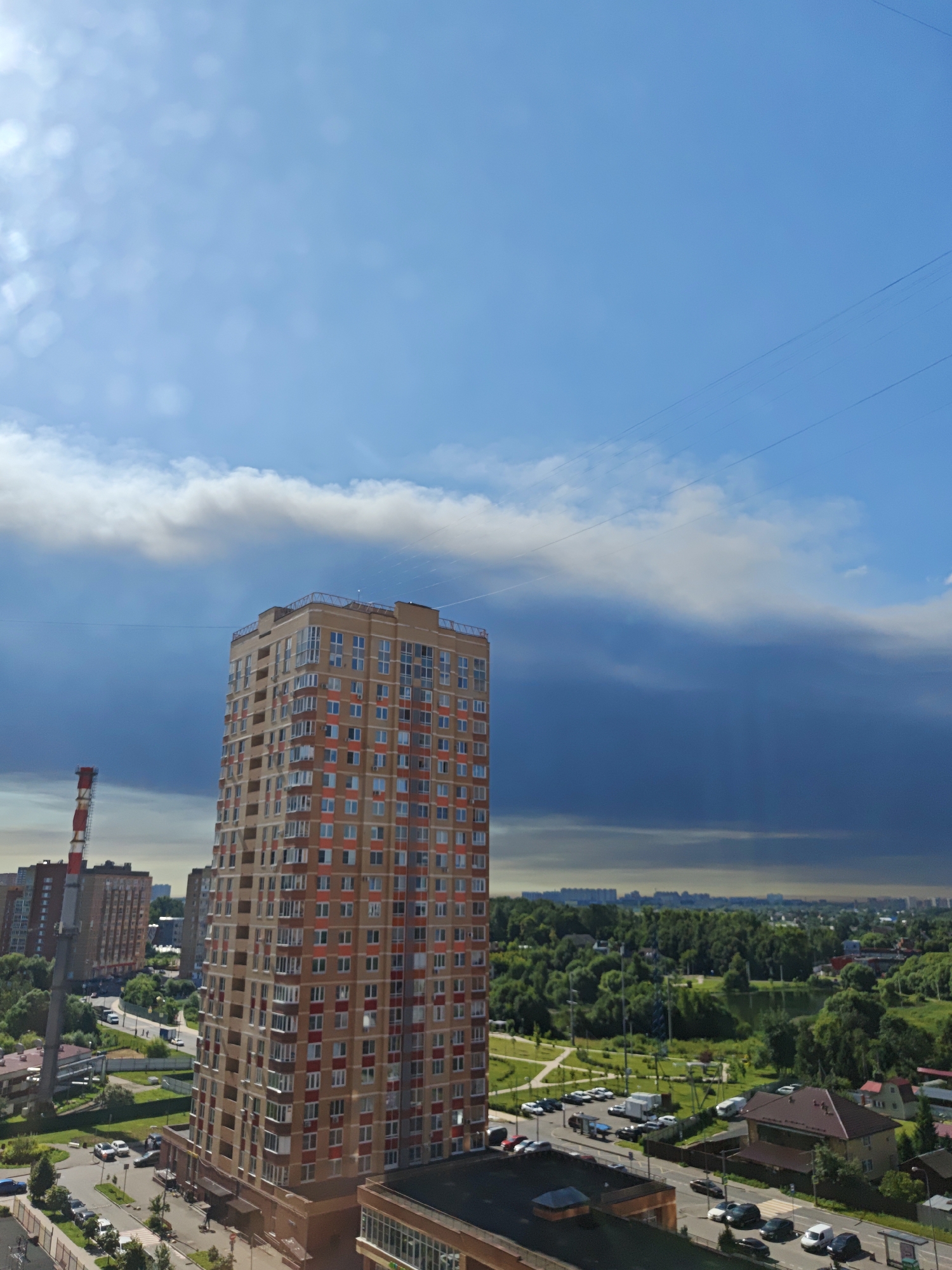 The image captures an urban landscape in Shcherbinka, Russia, dominated by a tall, multi-story residential building. The building, with a facade of light brown and orange, features numerous windows and balconies, indicative of a modern apartment complex. In the background, other buildings and a tall industrial chimney with red and white markings are visible, suggesting a mix of residential and industrial areas. The scene is set against a dramatic sky, with a bright blue expanse above, contrasted by dark, heavy clouds that suggest an approaching storm or recent rainfall. The foreground includes a road with parked cars, a pedestrian crossing, and surrounding greenery, including trees and a body of water in the distance, hinting at a park or natural area within the city. The overall impression is of a dynamic urban environment under a changing sky.