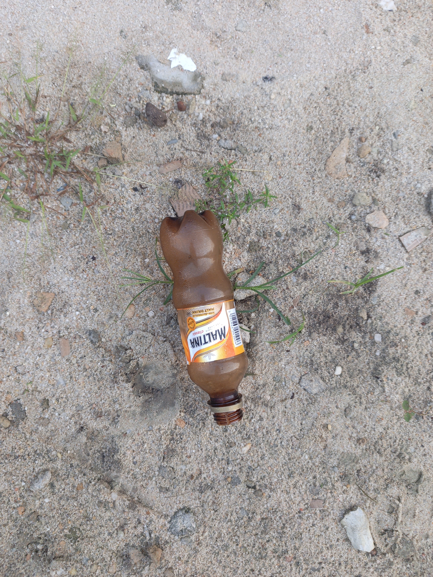 A brown Maltina bottle lies on a sandy surface interspersed with small rocks and sparse green vegetation. The bottle is positioned horizontally, with its label visible, slightly embedded in the sand. Above the bottle, there is a piece of white debris. The location is Unknown, Unknown.