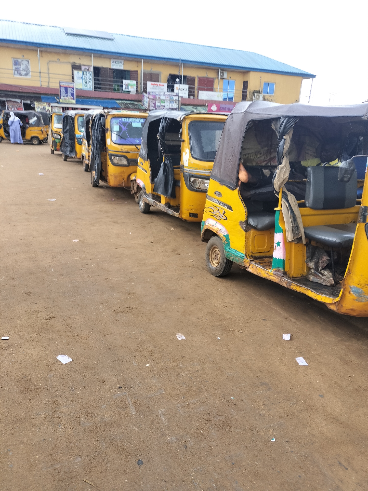 The image depicts a row of parked yellow "Keke Napep" auto-rickshaws in Ikorodu, Nigeria. These three-wheeled vehicles are lined up on a dirt or sandy ground. A building with a blue roof and visible signage, potentially indicating commercial establishments, is in the background. One person is walking nearby. Debris or small pieces of paper litter the ground.