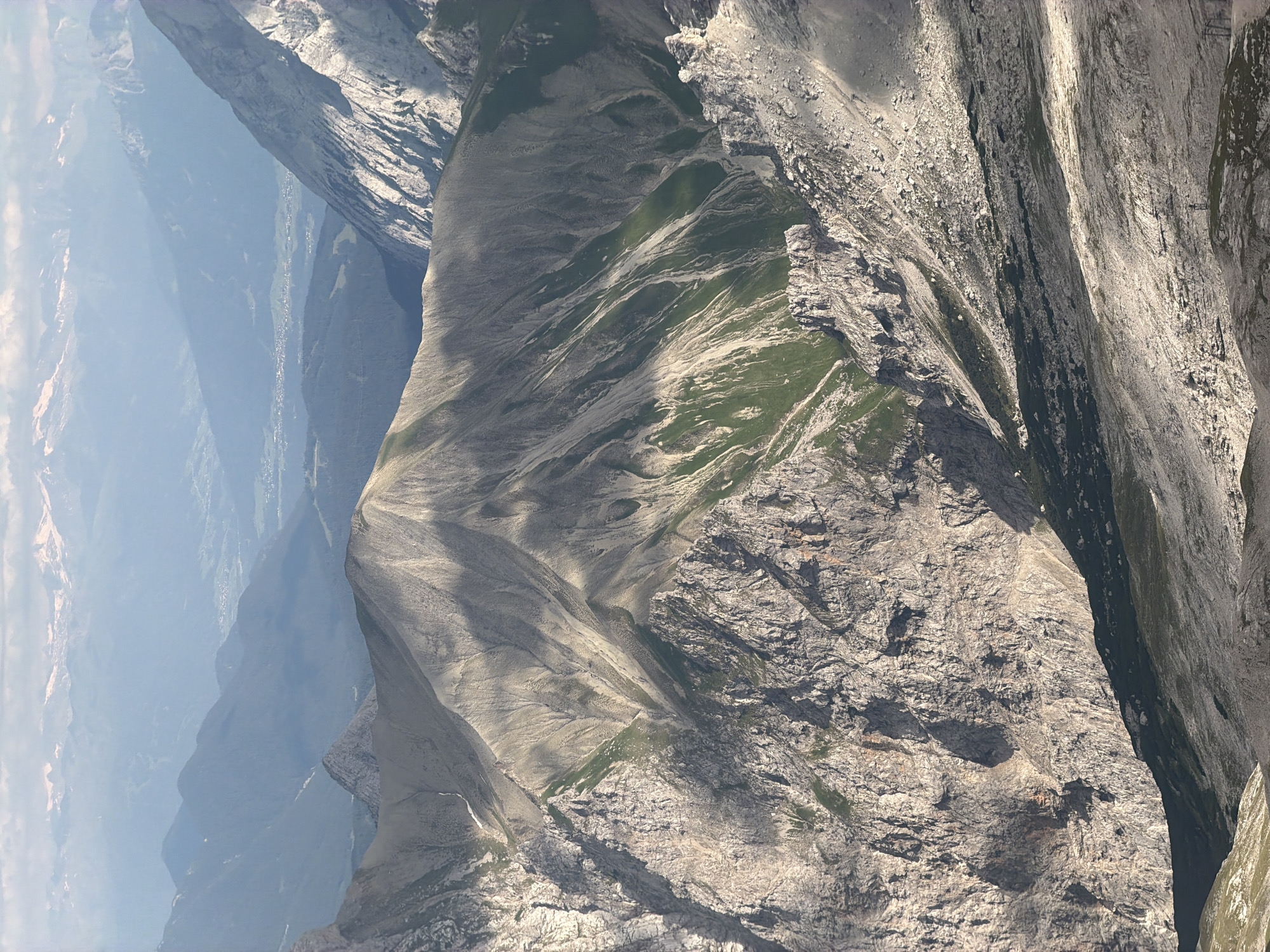 The image captures a dramatic, sun-drenched mountain landscape in Garmisch-Partenkirchen, Germany. The scene is dominated by rugged, rocky cliffs and steep mountain slopes, with patches of vibrant green grass interspersed across the terrain. Shadows play across the mountainside, indicating it is likely midday or early afternoon with clear, bright weather. The perspective is a high-angle shot, looking down into a deep valley or chasm formed by the imposing mountain ranges.
In the distance, a hazy view of a valley floor is visible, with what appear to be scattered buildings suggesting a settlement nestled within the mountainous terrain. Further back, softer, blue-hued mountain ranges recede into the horizon, creating a sense of immense scale and depth. The textures of the rock are detailed, showing the weathered and fractured surfaces characteristic of alpine environments. The bright sunlight accentuates the contours and sharp edges of the rock formations. No people or man-made objects are clearly discernible, emphasizing the wild and natural state of the location. The overall impression is one of grandeur, solitude, and the raw beauty of the natural world in a high mountain setting.