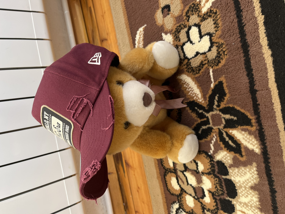 An indoor scene in Sheynovo, Bulgaria, depicts a light brown plush teddy bear positioned centrally on a patterned rug. The teddy bear wears a maroon baseball cap, which exhibits a distressed aesthetic with multiple tears on its brim and front panels. A rectangular patch on the cap's front reads "BUFFALO NY New Era EST. 1920," and a white embroidered New Era logo is visible on the cap's left side. A light brown ribbon is tied around the teddy bear's neck. The rug features a predominantly brown background with abstract floral or paisley patterns in black, cream, and lighter brown hues. In the background, a white horizontally ribbed radiator is situated above a light-colored wooden floor.