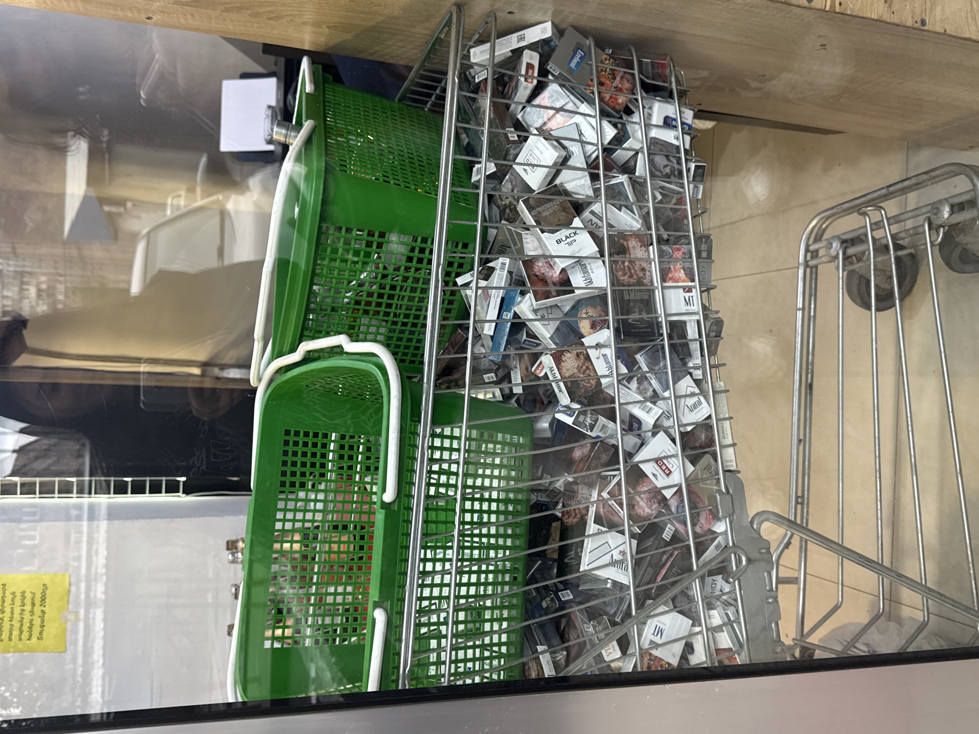 The image depicts a display case filled with cigarette packs in Yerevan, Armenia. Two green plastic baskets are positioned to the left of a wire rack overflowing with cigarette packs. Brands such as 'Ararat', 'Akhtamar', 'Black Tip', 'Dunhill', 'MT', and 'Red Line' are visible among the assortment. A yellow sign with Armenian text and the number 2000 is affixed to the display case. A metal trolley stands to the right.