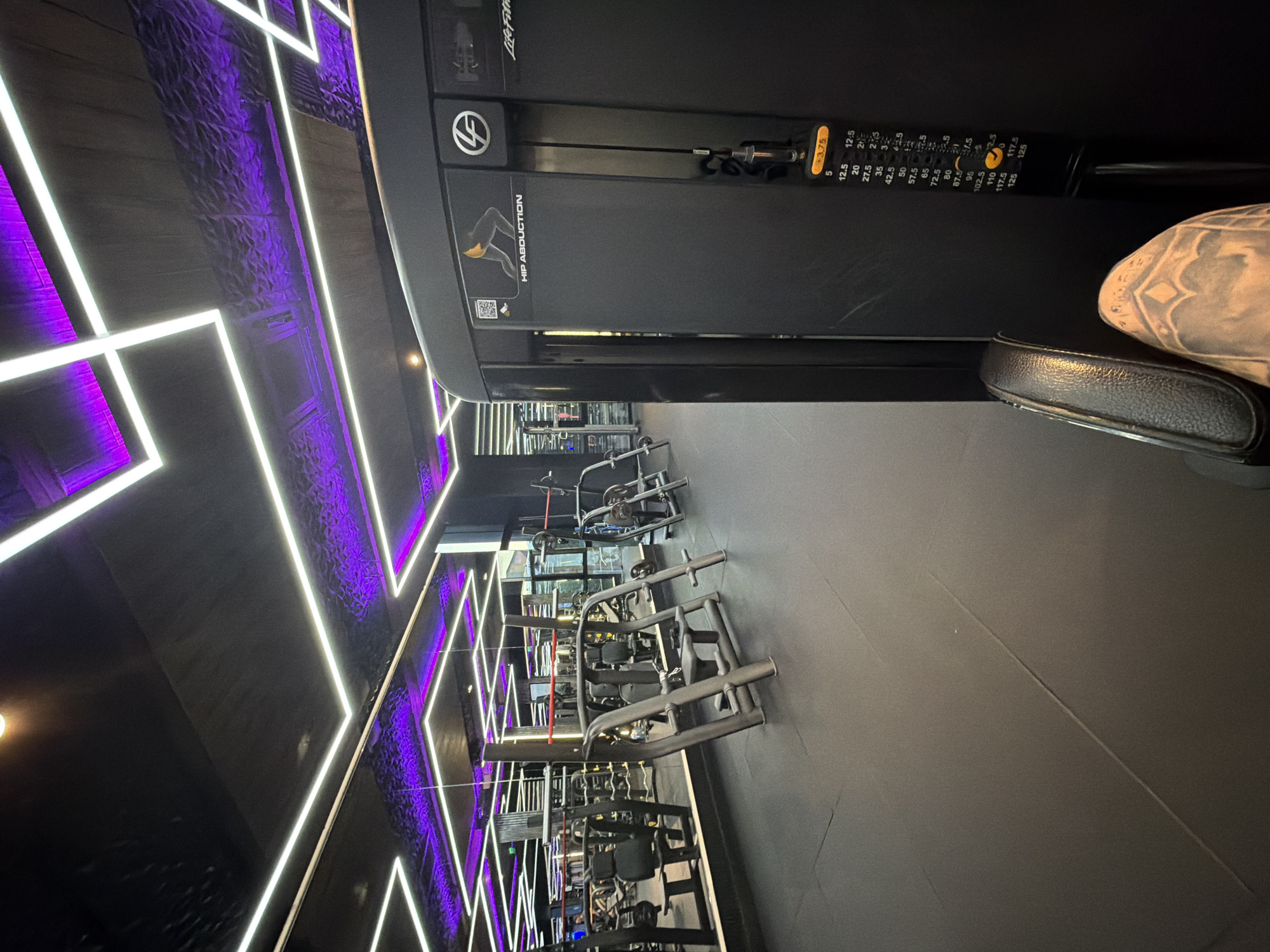 The image displays a modern gym interior in Plovdiv, Bulgaria, devoid of people. In the foreground, a black LifeFitness "Hip Abduction" machine is clearly visible. It features a padded seat and a weight stack showing various resistance increments, including "5", "12.5", "20", "27.5", "35", "42.5", "50", "57.5", "65", "72.5", "80", "87.5", "95", "102.5", "110", "117.5", and "125", along with a smaller "+3.75" weight.
The gym boasts a distinctive design with dark walls and ceiling illuminated by vibrant purple and white LED strip lights arranged in geometric patterns. These lights create a dynamic and energetic ambiance. Further into the background, an array of other gym equipment, including various strength training machines, can be seen, lined up along the dark, smooth floor. The overall impression is of a stylish and well-equipped fitness facility.