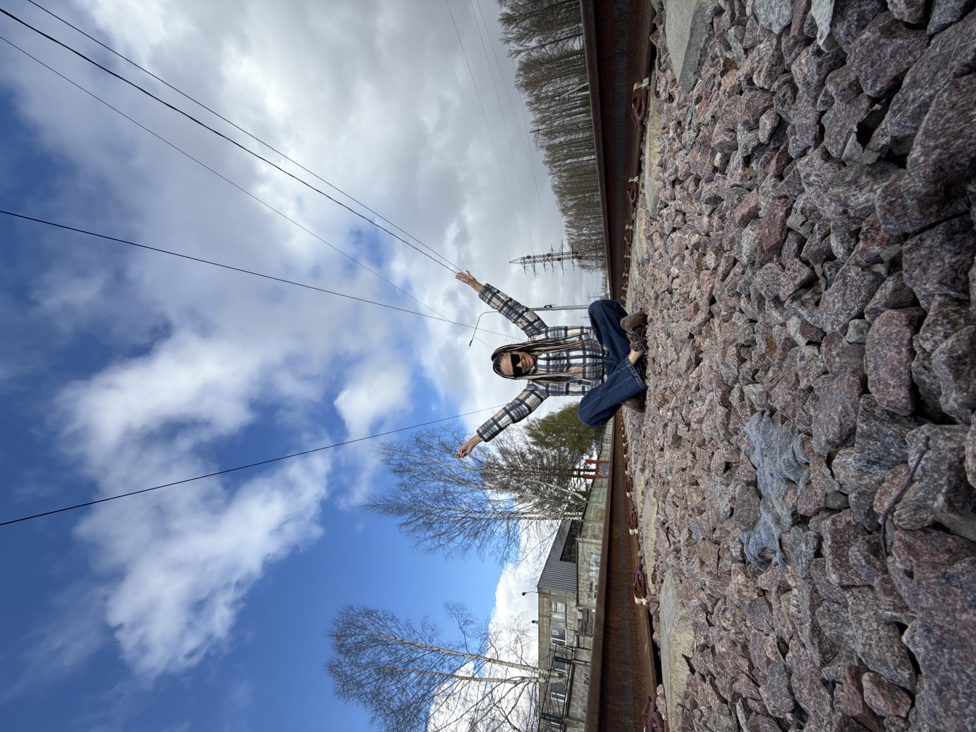 A person is depicted sitting on a railway track, posing with outstretched arms. The original image is rotated 90 degrees clockwise, making the railway ballast appear vertical.
The main subject is an individual with dark dreadlocks or braids, wearing sunglasses, a plaid shirt with black, white, and possibly blue checks, and blue jeans. They are wearing dark brown hiking boots. A thin wire, possibly from headphones, extends from their right hand.
The setting is an outdoor railway environment. The person is seated on the edge of the track, composed of rusty metal rails, wooden sleepers, and crushed stone ballast. In the background, power lines stretch across a partially cloudy sky, which shows patches of blue. Bare deciduous trees are visible, suggesting a late winter or early spring season. A line of dense conifer trees and a tall metal electricity pylon are in the distant background on the right. To the left of the tracks, there are buildings, including what appears to be an industrial structure or warehouse.
The activity is a posed photograph, with the individual adopting an expansive stance. The time of day is daytime, under natural light. The weather is partly cloudy. No legible text is visible in the image. The location context provided is Rusanovo, Russia.