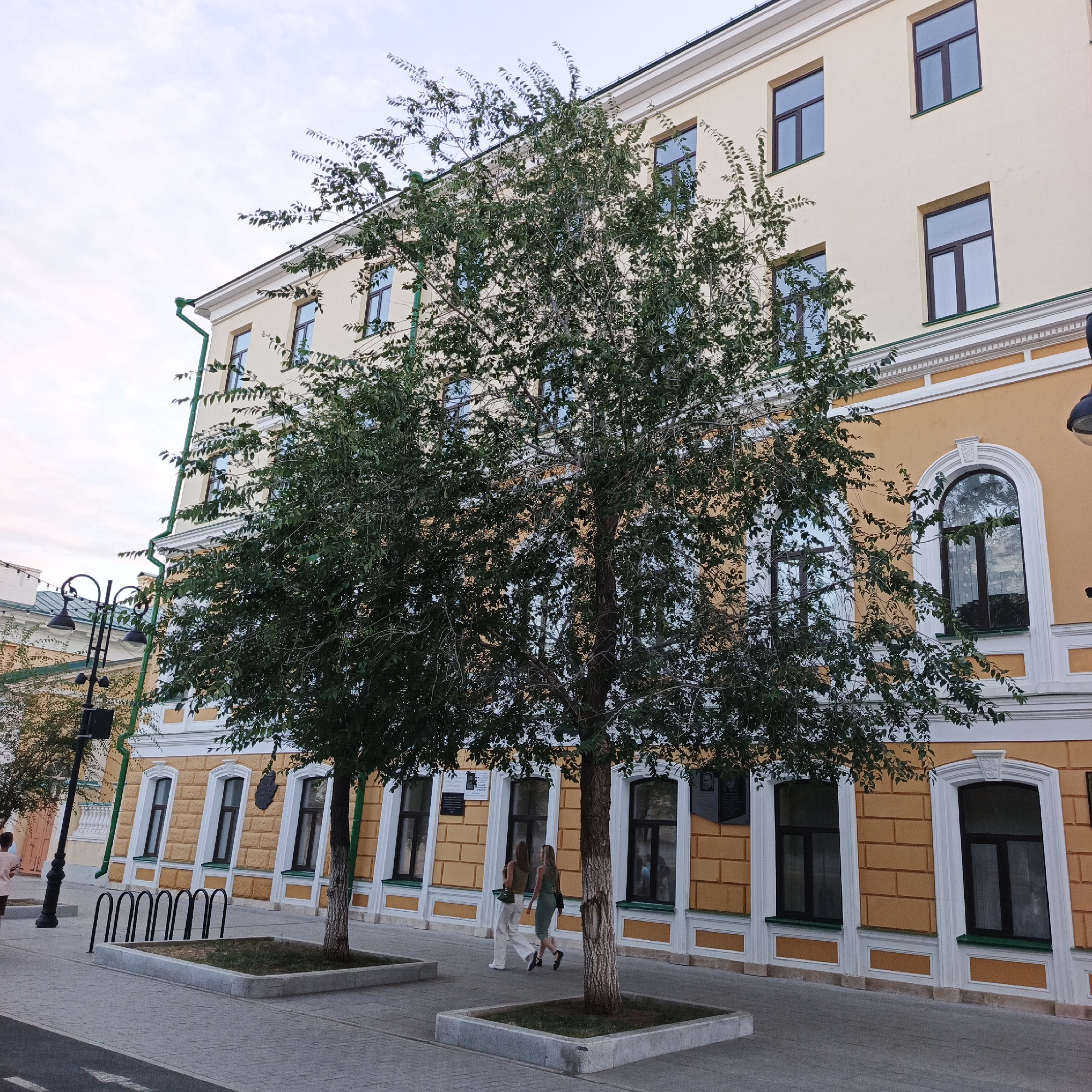 This image depicts a street scene in the city of Orenburg, Russia. In the foreground, a paved sidewalk runs alongside a building. Two young women are walking on the sidewalk, heading away from the camera. The building behind them is a multi-story structure with a yellow facade, white trim, and dark-framed windows. The lower portion of the building features an ornate brick pattern. Two young trees are planted in square planters on the sidewalk, partially obscuring the building. A black lamppost with three lights stands to the left of the trees. In the background, more of the building and a cloudy sky are visible.