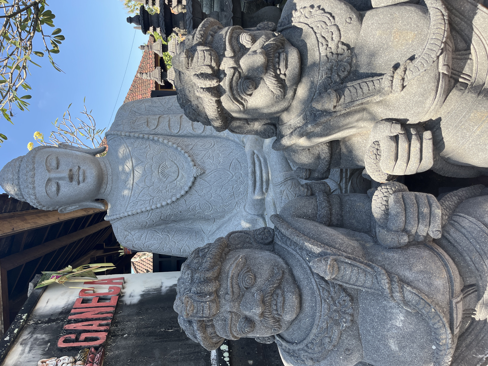 The image depicts a stone statue of Buddha in a seated meditation pose, with two smaller stone statues in the foreground. The statue of Buddha is carved from gray stone and wears a beaded necklace. The two foreground statues are of warrior figures with stern expressions, mustaches, and elaborate headdresses. They are also carved from gray stone and appear to be holding objects in their hands.
Behind the statues, there is a building with a wooden roof and tiled roof. To the left of the Buddha statue, a red sign with the word "GANECA" is visible on a weathered wall. The environment appears to be outdoors, with a clear blue sky and some green foliage visible.
This scene is located in the city of Sukawati, Indonesia. Sukawati is known for its art markets and traditional crafts, making it a likely location for such stone carvings. The overall atmosphere is serene, with the statues suggesting a spiritual or cultural site.