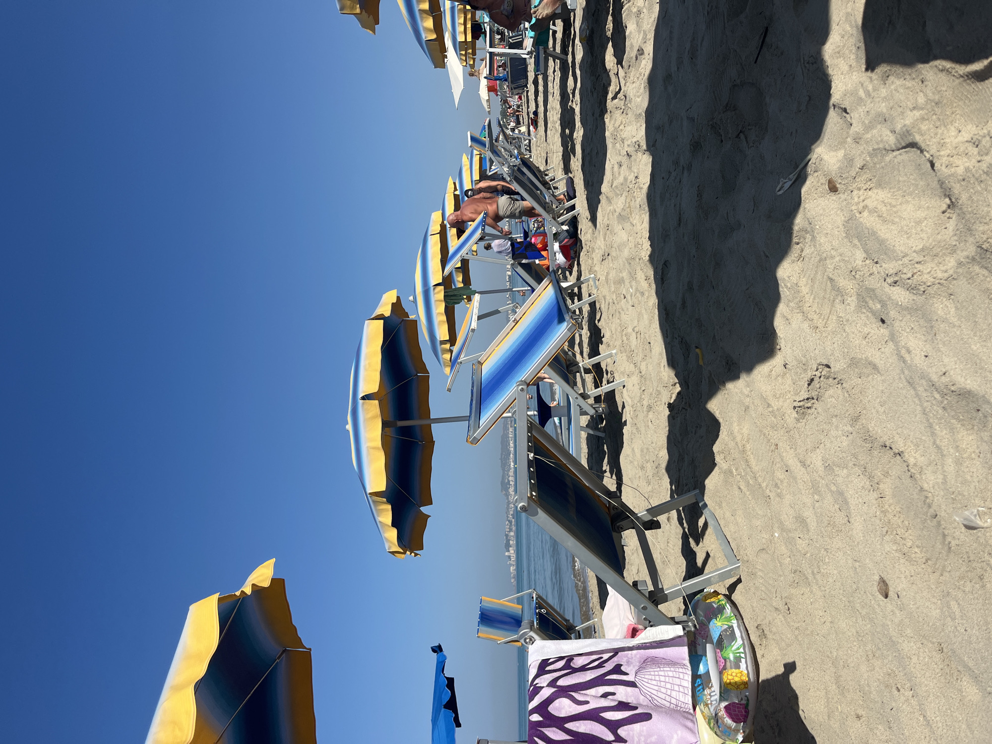 The image depicts a crowded beach scene on a clear, sunny day. In the foreground, several beach chairs are set up on the sand, some with colorful blue and yellow striped upholstery and matching umbrellas. A purple towel with a white coral pattern and a small inflatable ring with a pineapple design are draped over one of the chairs. The sea is visible in the background, with a faint city skyline in the distance. Several people are scattered across the beach, some standing and others sitting in chairs, while others are visible wading in the shallow water or further out in the sea. The overall impression is one of a popular vacation spot.