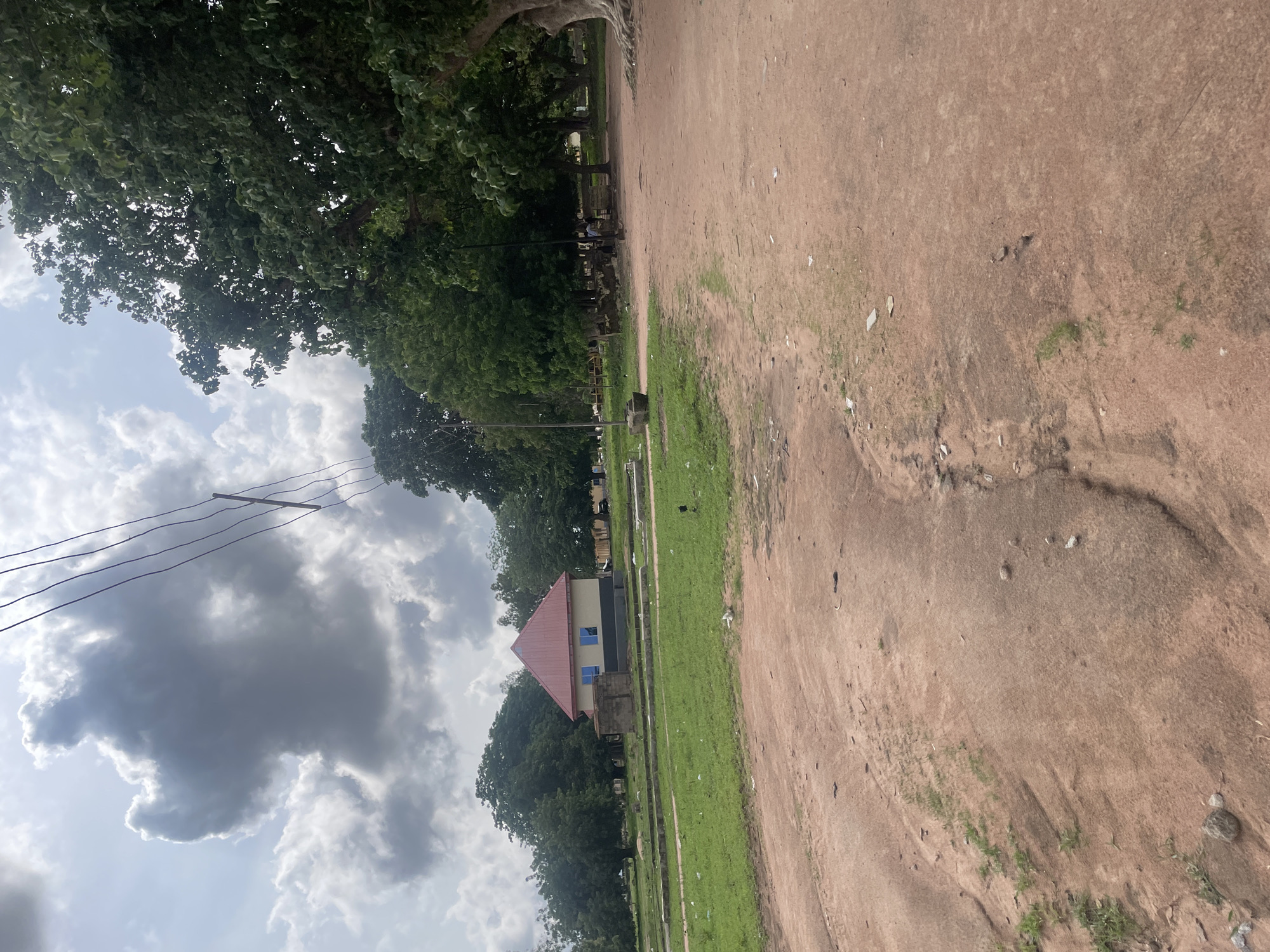 The image depicts an outdoor scene with a dirt path in the foreground, leading towards a grassy area with scattered debris. A small, light-colored building with a red, gabled roof and blue windows is visible in the mid-ground. Several large trees with green foliage are present, with one particularly large tree on the right side of the frame. Power lines are strung across the upper portion of the image. The sky is partly cloudy with dramatic, dark clouds. A few indistinct figures can be seen in the distance near some structures under the trees. The overall impression is of a rural or semi-urban environment under a partly overcast sky.