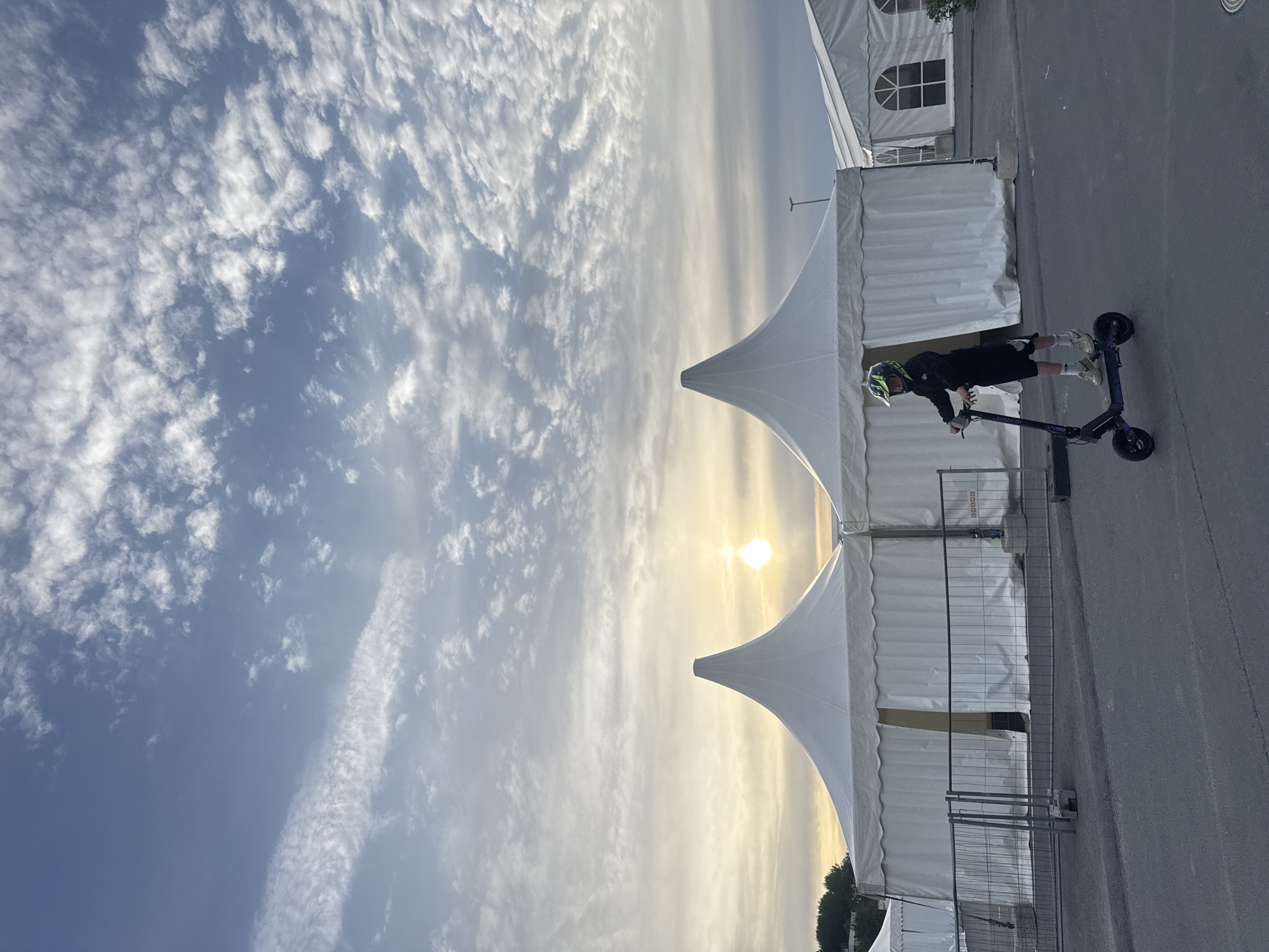 A young person wearing a helmet rides a scooter in front of two large white tents. The tents have conical roofs and are set up on a paved area. The sky above is filled with clouds and the sun is visible, appearing as a bright orb. The scene suggests an outdoor event or venue. The location is not specified as Tallinn, Estonia.