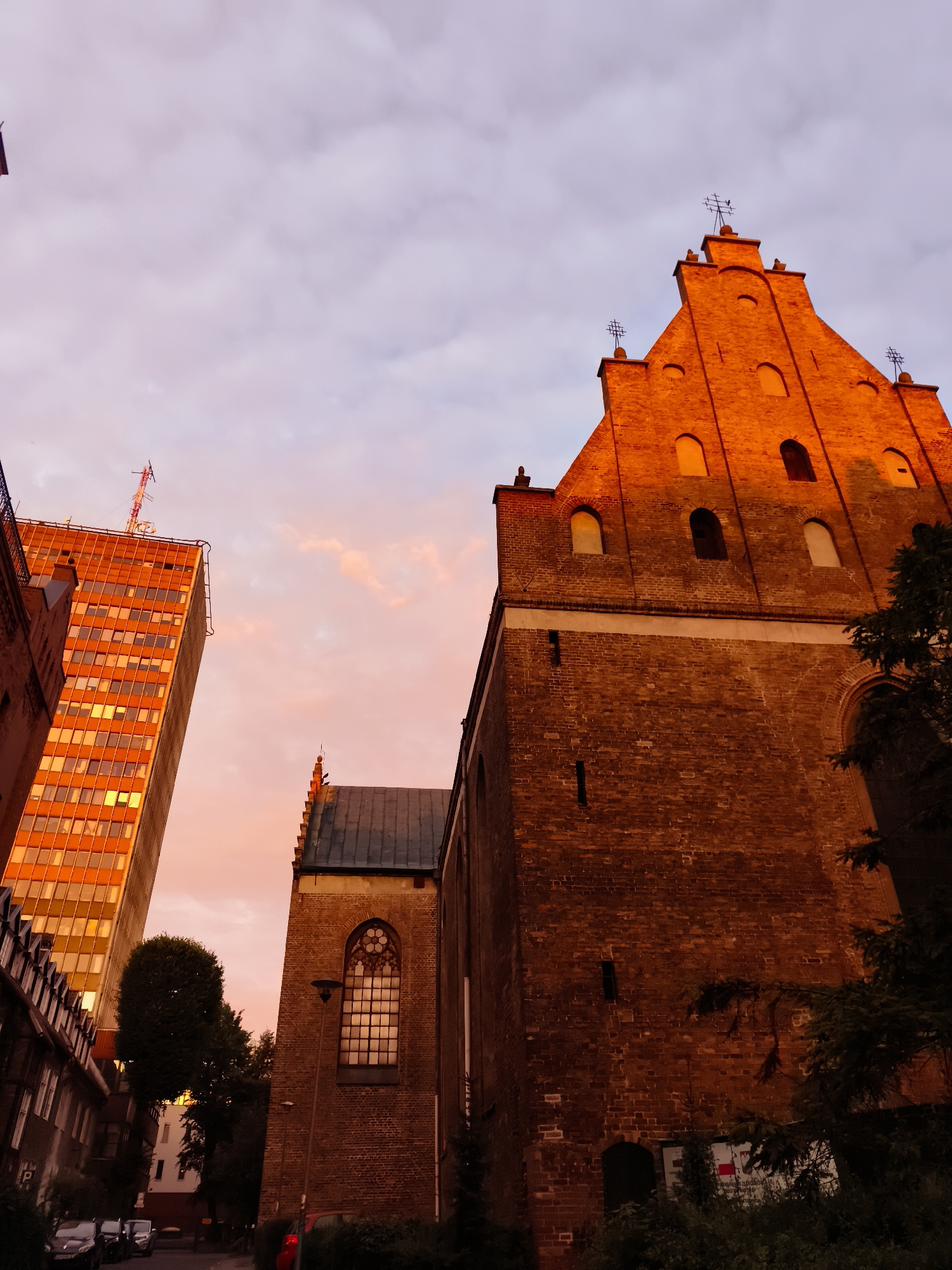 The image shows a low-angle view of a brick church and a modern skyscraper against a sky with pink and orange hues. The church, with its steep roof and Gothic-style windows, is bathed in the warm light of sunset. The skyscraper, to the left, has a facade of orange-tinted glass and a radio mast on top. The scene appears to be set in an urban environment, possibly in Gdańsk, Poland, given the architectural styles and the presence of cars parked on a street in the foreground. The overall impression is one of a juxtaposition of old and new architecture, captured during a vibrant sunset.
