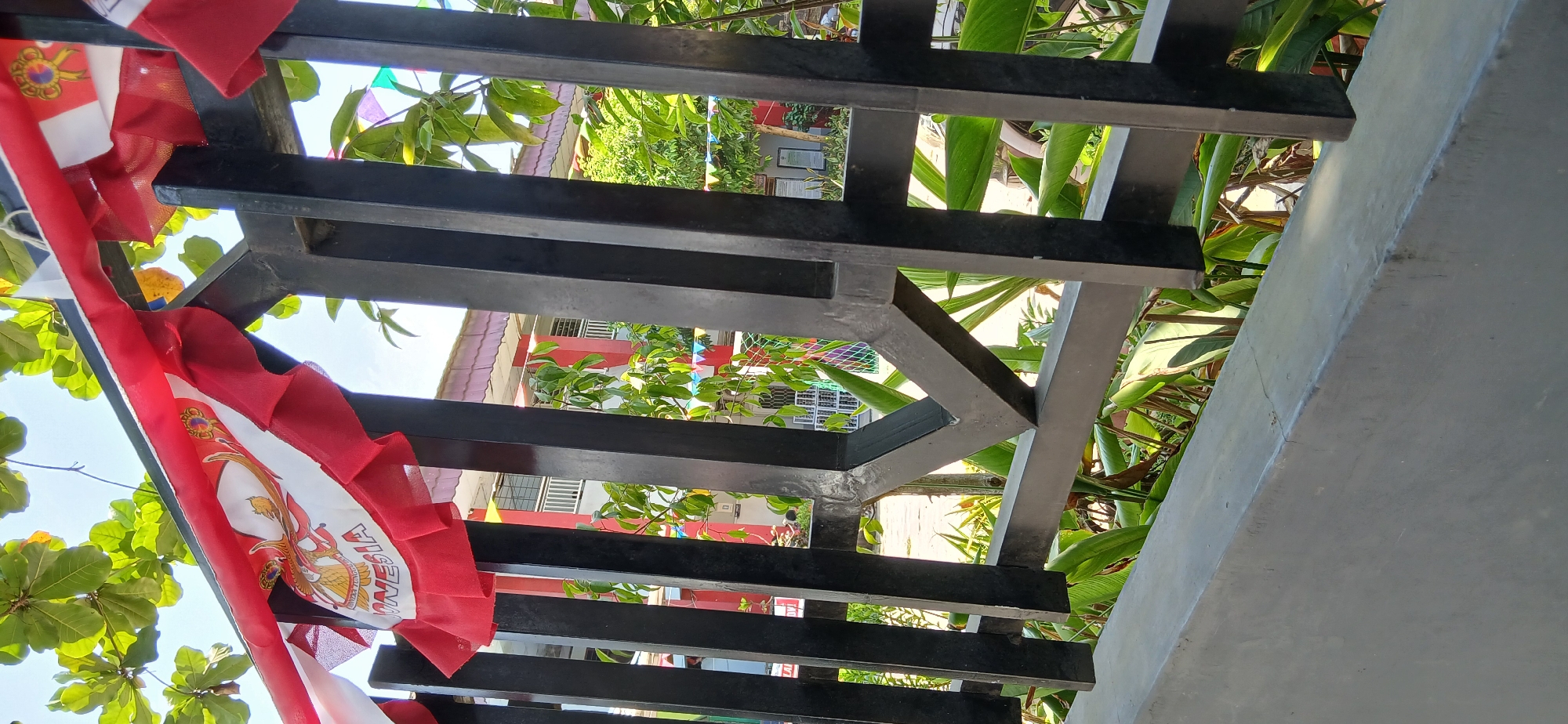The image depicts a section of a black metal fence with decorative elements and Indonesian flags draped across the top. Behind the fence, lush green foliage is visible, interspersed with glimpses of a building with red and white accents. The building appears to be a school or similar institution, with windows and a tiled roof. Small, colorful flags are strung along the fence further down. The overall scene suggests an outdoor setting, possibly a schoolyard or a public space decorated for a national celebration. There are no people visible in the image, and the focus is on the fence, flags, and surrounding environment. The image does not provide any specific location within Samarinda, Indonesia.