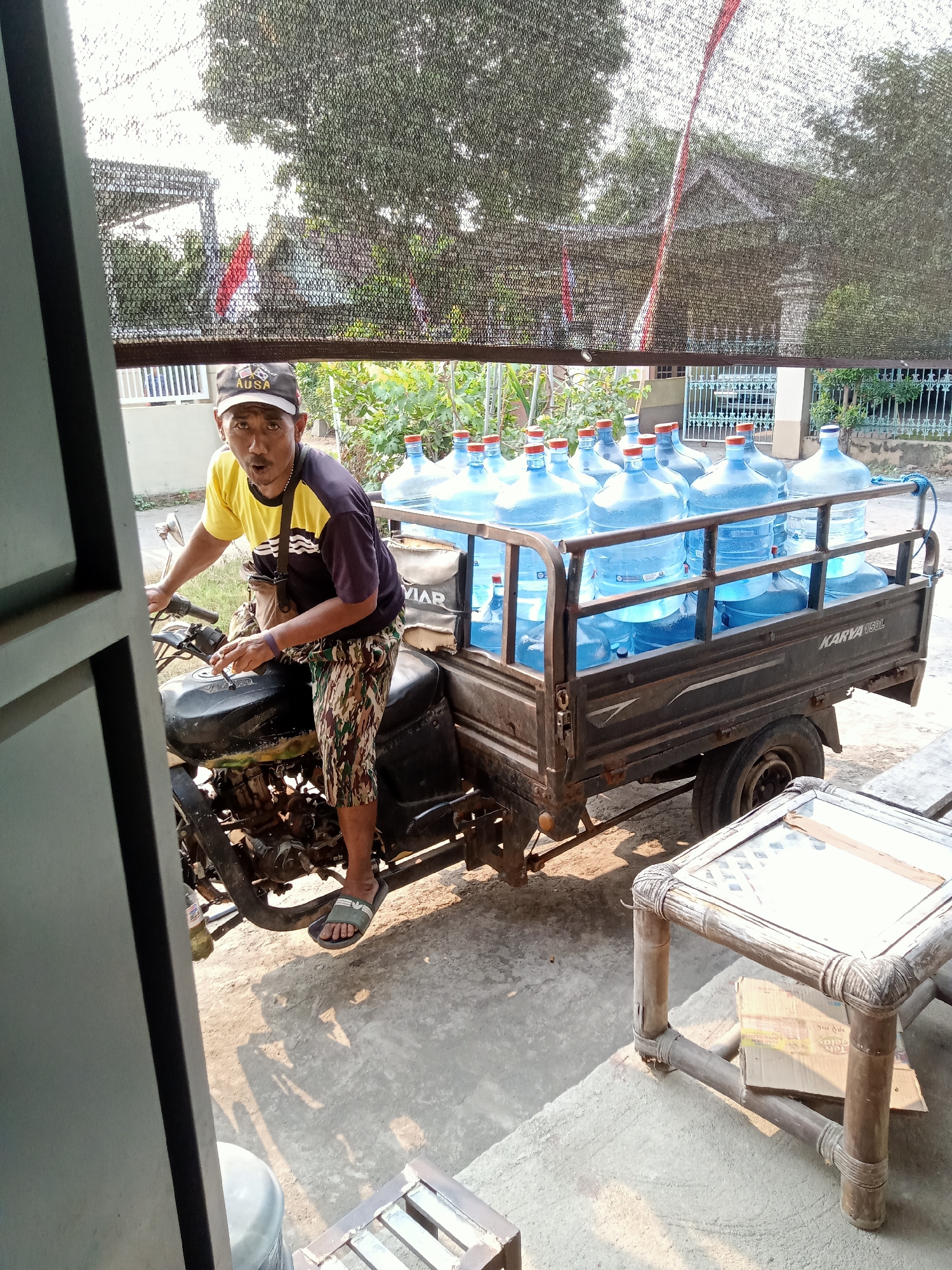 In Simo, Indonesia, a man wearing a yellow and dark blue t-shirt, camouflage shorts, and a baseball cap is seated on a black three-wheeled motorcycle with a trailer. The trailer is filled with multiple large, clear plastic water jugs with red caps. The man appears to be looking directly at the camera, with his mouth slightly open. The motorcycle is parked on a concrete surface, and there is a rustic wooden table with a glass top to the right of the trailer. In the background, a fence and trees are visible, with a hint of a building and Indonesian flags. A black net-like covering is stretched across the upper part of the scene, possibly for shade.