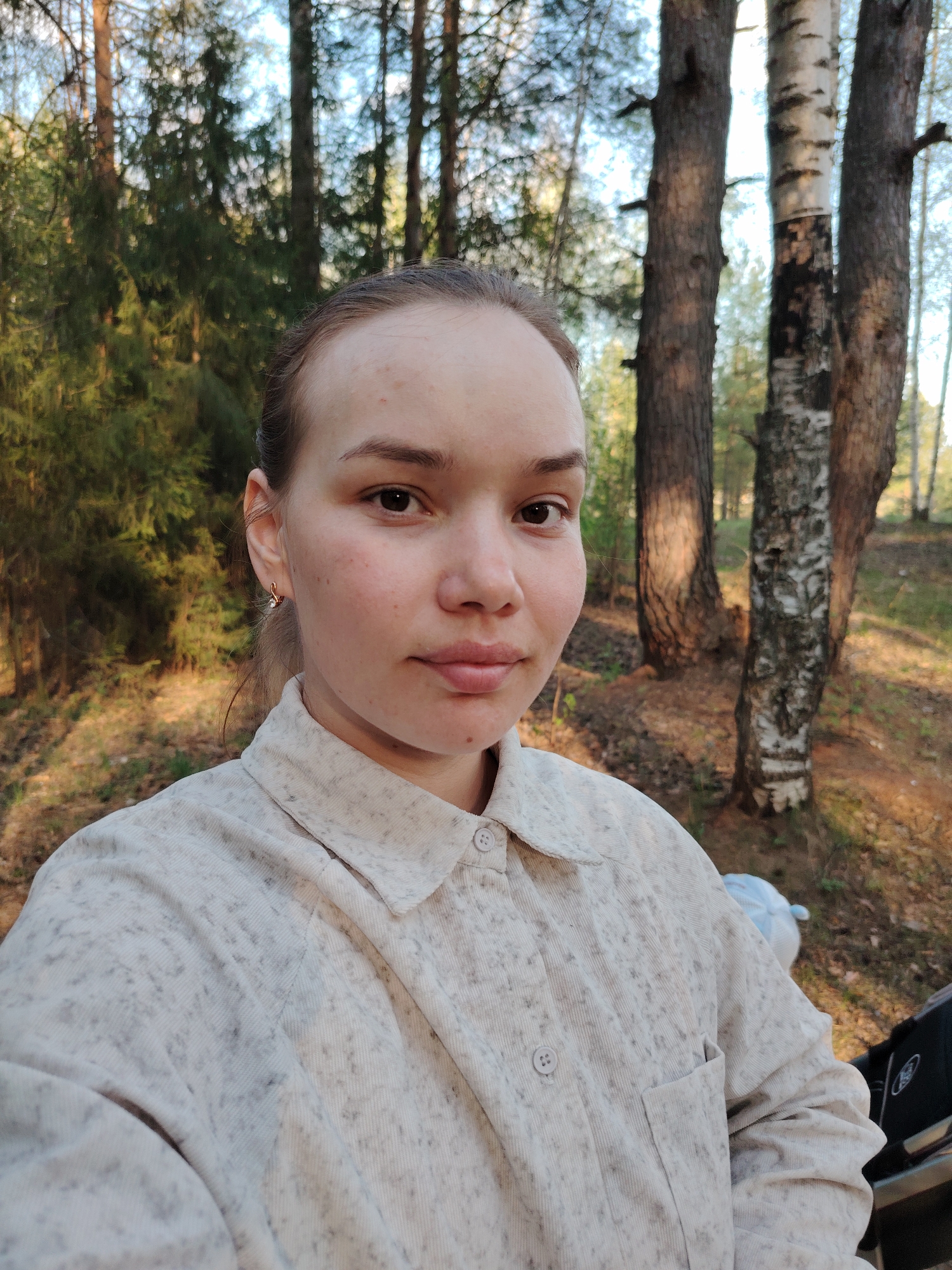 A woman is standing outside in a forest of pine and birch trees. She is wearing a light-colored shirt with a collar and a pocket. The background includes trees and a dirt path. Based on metadata, the location is near Марковский, Russia.