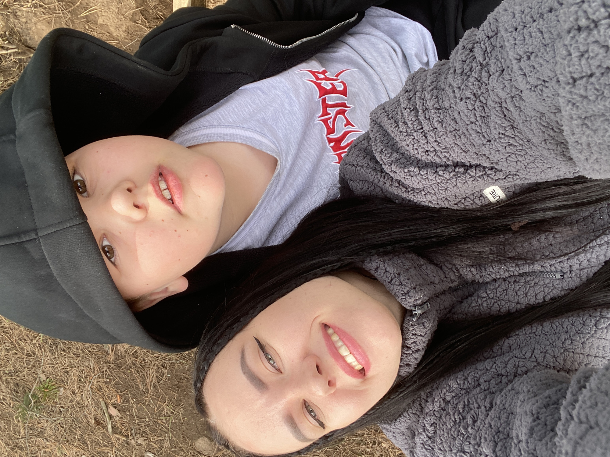 An overhead photograph captures two individuals, likely young women, lying side-by-side on the ground outdoors, looking up at the camera. The setting is a natural environment, with the ground covered in dry pine needles, small twigs, and patches of earth, indicative of a wooded or forest area in Chaikovsky, Russia. The lighting suggests it is daytime.
The person on the left has fair skin, light brown eyes, and freckles. They are wearing a black hooded sweatshirt over a light grey t-shirt that features a prominent red, stylized graphic resembling text on the chest. Their expression is a gentle, natural smile.
The person on the right has fair skin, long dark hair, which includes visible braids. They are wearing a grey, textured, fuzzy sherpa-style jacket with a zipper. A small white fabric tag on the jacket's sleeve is partially visible, showing text that appears to be "PURE". This individual has noticeable winged eyeliner and pink lipstick, and displays a wider smile with teeth visible.
Both individuals appear relaxed and are posing for the photograph, conveying a casual and friendly mood.