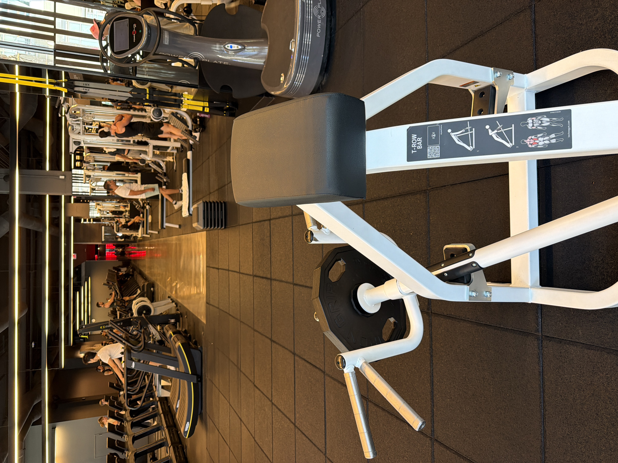 The image captures a gym scene in Los Angeles, United States. The foreground features a T-row bar machine with a black seat cushion and a weight plate attached. The machine has white framework and instructional diagrams. In the background, gym-goers are using various exercise equipment, including rowing machines and treadmills. The gym has a modern aesthetic with dark flooring and overhead lighting.