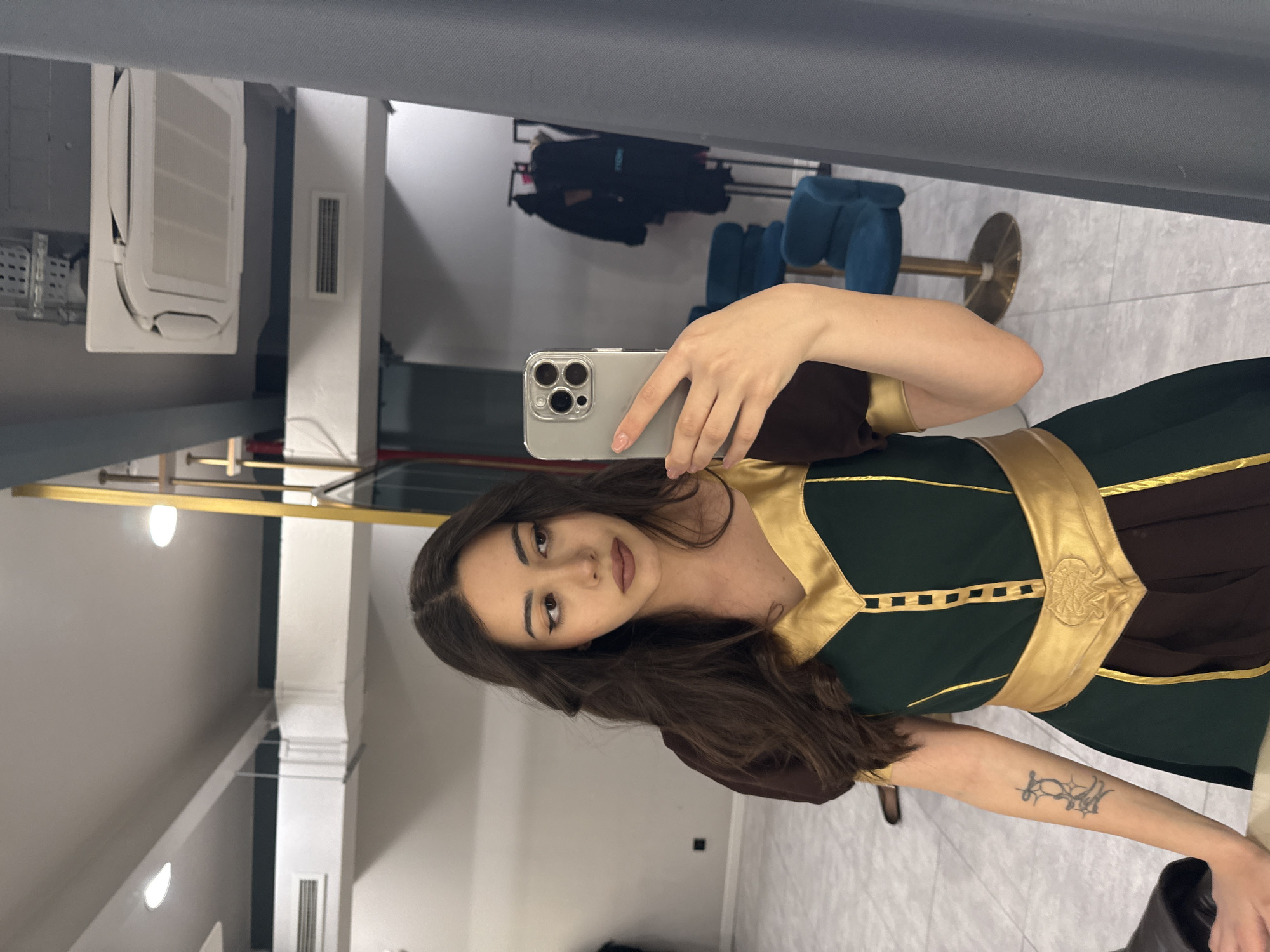 A woman in a mirror selfie indoors. She is wearing a brown and green dress with gold accents. She is holding a smartphone in her left hand. The background appears to be a retail space, possibly a clothing store, with clothing racks and blue chairs visible in the mirror. The ceiling and walls are white. There is a vent and an air conditioning unit in the upper left. The specific location is difficult to determine from the photo, and there is nothing to indicate it is in Yerevan, Armenia.