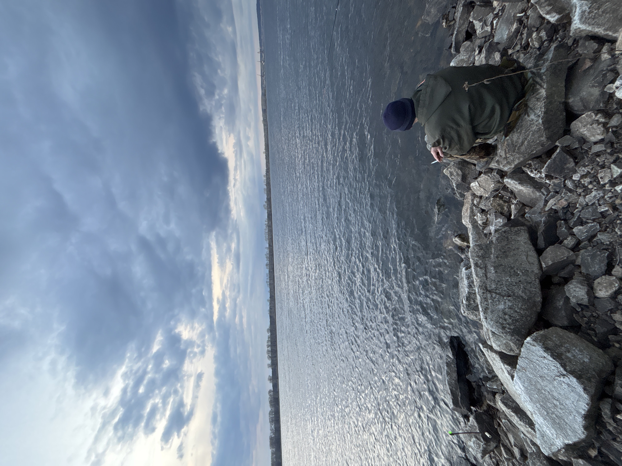 The image depicts a solitary person on a rocky shoreline beside a large body of water under a cloudy sky. The person, seen from behind, is seated on large grey rocks at the water's edge. They are wearing a dark blue knit hat and a dark green jacket. In their right hand, they hold a lit cigarette.
To the left of the person, a slender, green object, possibly a fishing rod or stick, is visible upright in the water, suggesting fishing as the activity. The vast expanse of water, likely a river or lake in Chaikovsky, Russia, stretches across the mid-ground, its surface showing ripples and reflecting the diffused light from the sky. A faint line of trees or distant land is discernible on the horizon across the water. The sky above is largely covered by dense, textured grey and white clouds, indicating an overcast day. No visible text is present in the image. The scene conveys a sense of quiet contemplation or peaceful solitude.