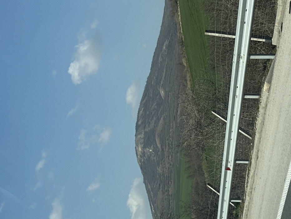 The image displays a rural landscape near Nevsha, Bulgaria, under a clear blue sky with scattered white cumulus clouds. In the immediate foreground, a segment of a paved road with a white demarcation line and an adjacent concrete curb is visible at the bottom left. A silver-colored metal guardrail, supported by vertical grey posts, extends horizontally across the lower middle of the frame, featuring a single red reflective marker. Behind the guardrail, a wire fence, supported by both wooden and metal posts, is partially obscured by a dense cluster of leafless, thorny bushes and small trees.
The midground consists of expansive, vibrant green agricultural fields, which slope gently upwards from the right side of the frame towards the background. These fields contrast with patches of drier, brown vegetation. Dominating the background is a large, rounded mountain. Its slopes are largely covered with sparse, dormant deciduous trees and shrubs, appearing in various shades of brown and grey. Lighter, possibly eroded or rocky, areas are visible on the upper portions of the mountain. No individuals or discernible actions are present in the scene.