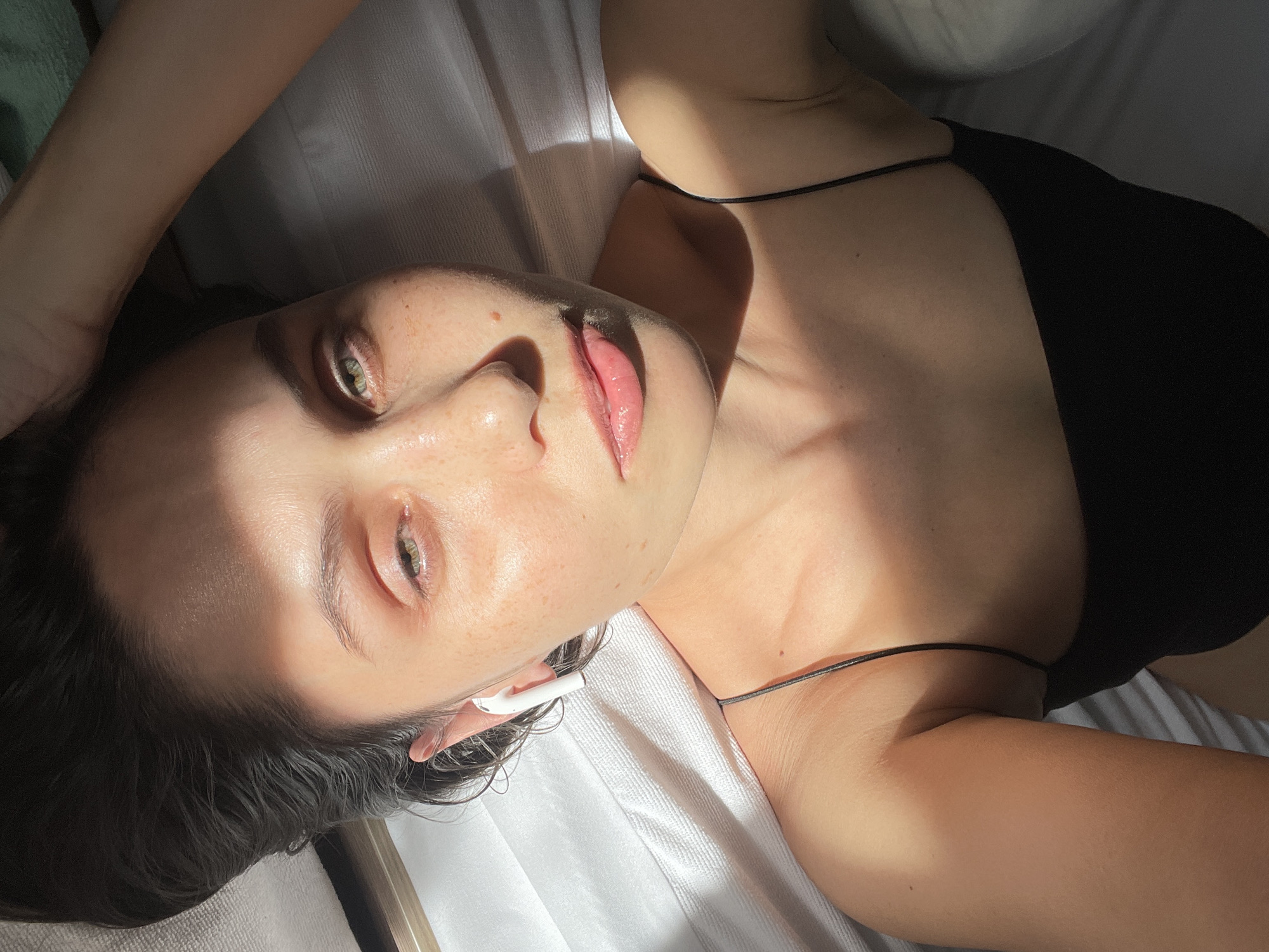 A light-skinned young adult female is lying on her back on a white sheet or bedspread. She is wearing a black top with thin straps. Her dark hair is wet and partially obscures her forehead. She has light green eyes and appears to have freckles. She is wearing an earbud in her left ear. The image is well-lit, with strong shadows across her face and body. The location is not identifiable from the image.