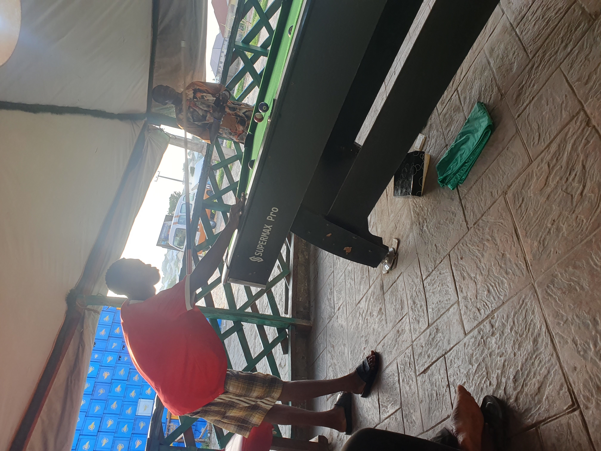 In Lokoja, Nigeria, two individuals are engaged in a game of pool at an outdoor establishment. The foreground is dominated by a pool table with a green felt surface, displaying several pool balls, including a red and a blue striped ball. The pool table has "SUPERMAX Pro" written on its side. To the left, a person wearing a red t-shirt and checkered shorts stands in profile, leaning over the table, appearing to be in the process of taking a shot. They are wearing black slides. To the right, another individual, dressed in a patterned shirt, stands near the end of the pool table, holding something in their hands. The ground is paved with textured square tiles. In the background, a green latticework fence is visible, beyond which are stacks of blue crates and some vehicles, including a white bus. The scene is partially covered by a beige awning. A folded green cloth and a tissue box sit on the tiled floor near the pool table. The lighting suggests it is daytime, possibly late afternoon, with sunlight casting shadows.