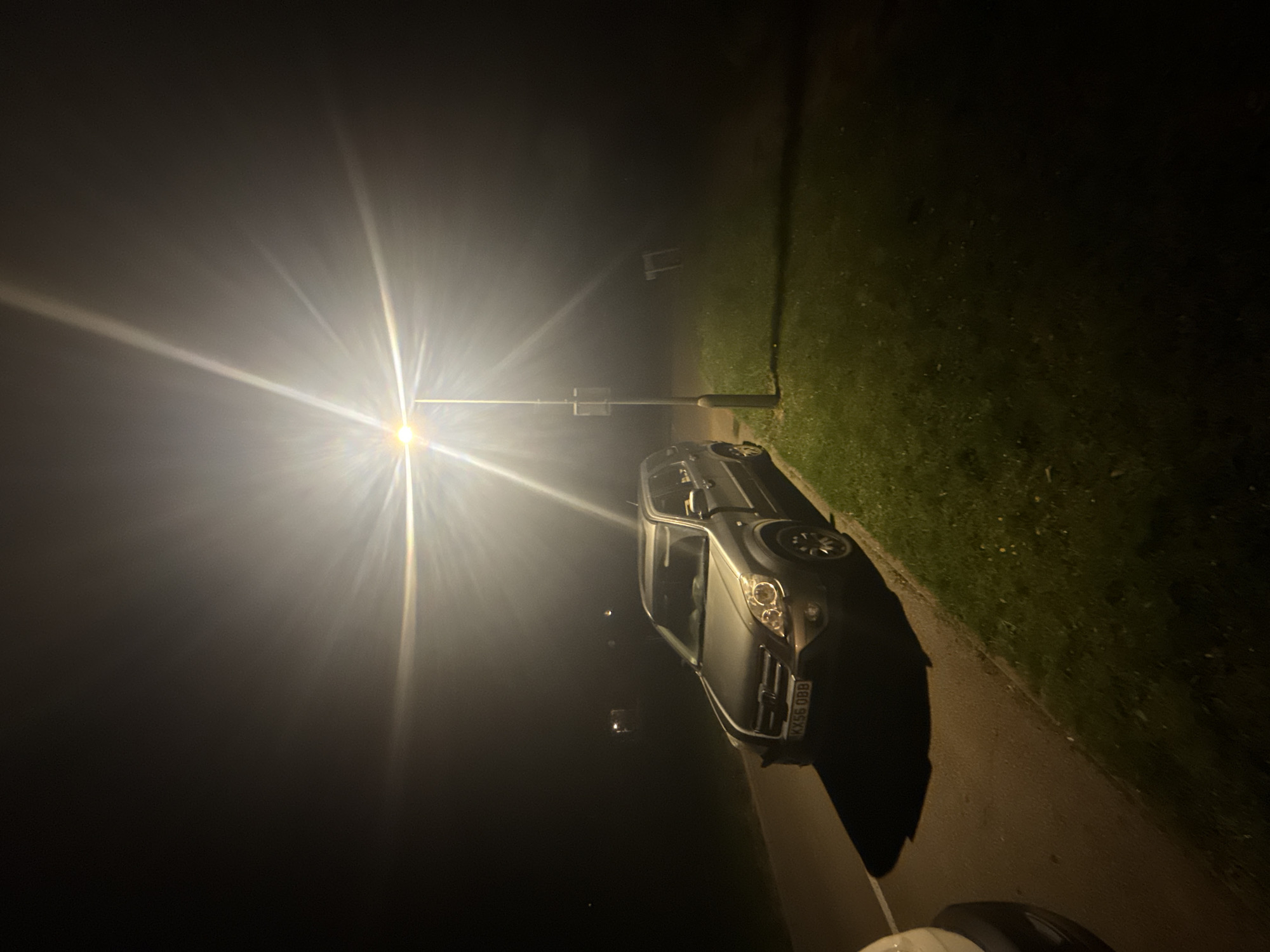 A nighttime scene in Mid Suffolk, United Kingdom. A silver Vauxhall Meriva (license plate KX56 OBB) is parked on a paved area next to a grassy verge. A street light illuminates the scene, creating a strong glare and long shadow. The background is dark. A white moped is in the bottom right corner.