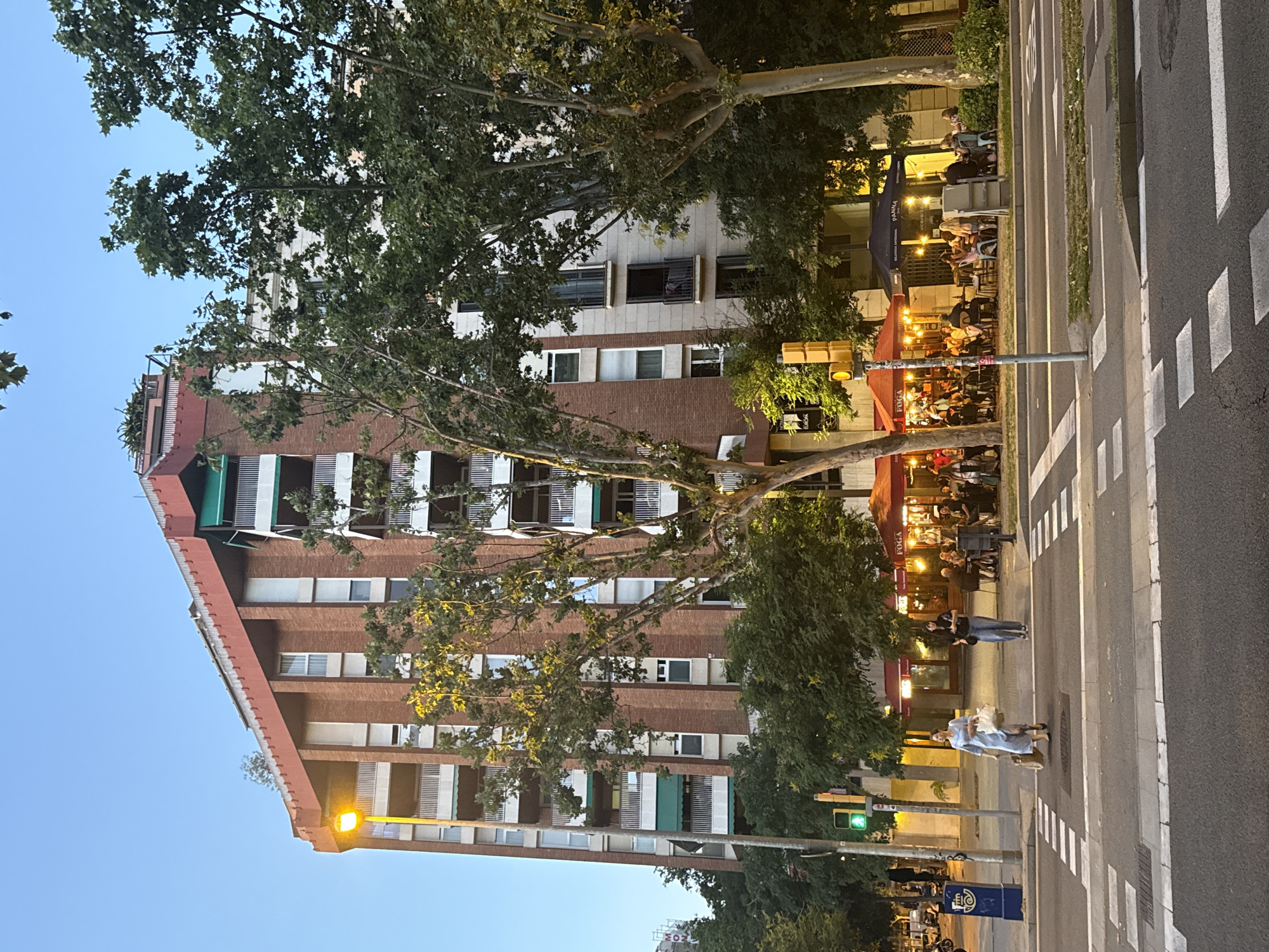 This is a daytime exterior shot of a building in Barcelona, Spain, with a street and sidewalk in the foreground. The building is tall, made of brick and light-colored stone, with multiple balconies and windows. Several large trees are in front of the building, some of which obscure parts of it. A street light illuminates the scene. On the sidewalk, there are a few people walking and some outdoor seating for a restaurant with umbrellas. A crosswalk is visible in the foreground, with two people crossing the street. The sky is a clear blue, suggesting it is a pleasant day.