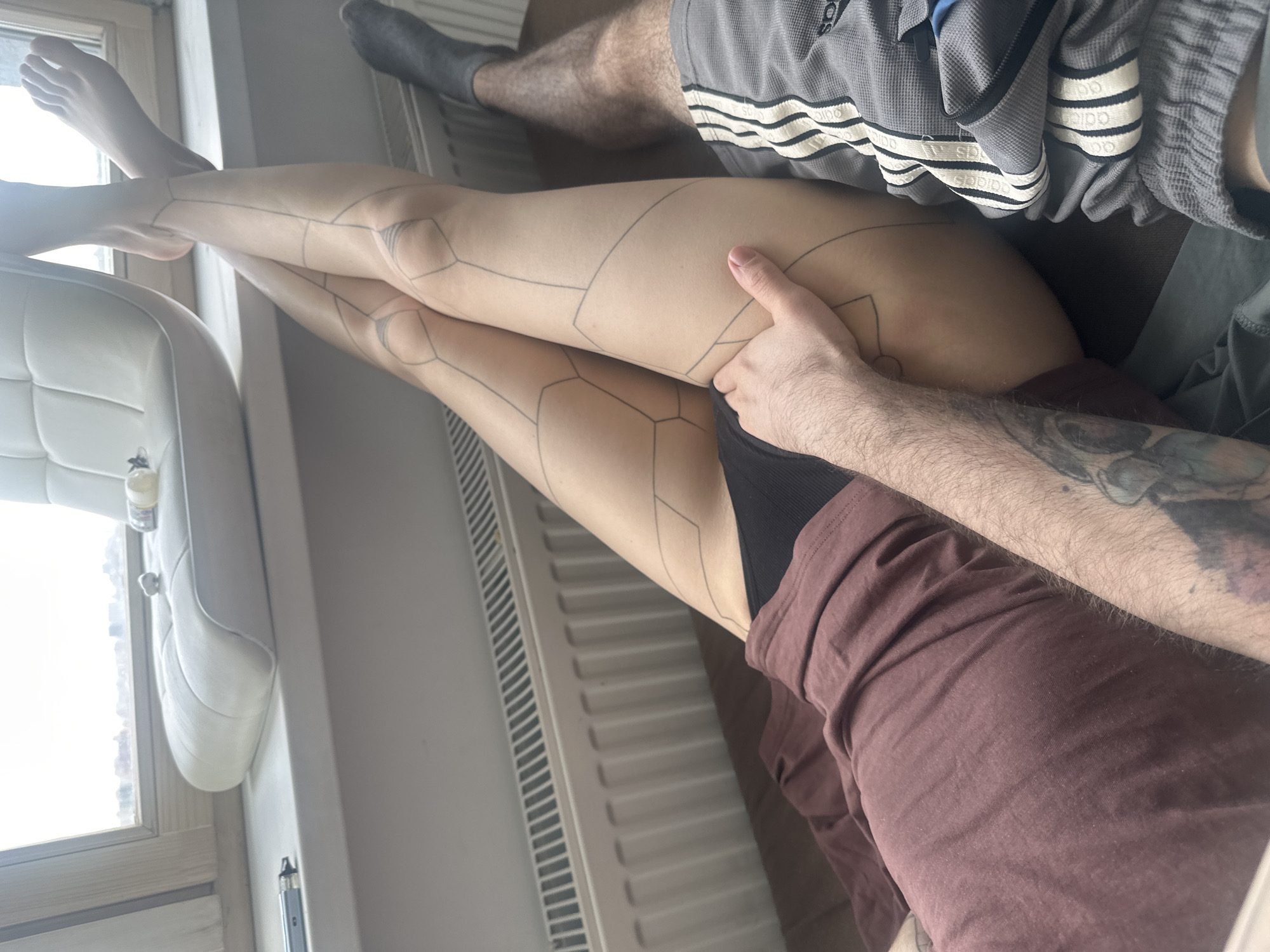 A person's legs are crossed and elevated, with geometric patterns resembling tattoo outlines visible on the skin. The person is wearing dark underwear. A second person is seated to the side, wearing brown clothing. An arm with a tattoo is visible, with a hand on the legs. Another person is wearing grey Adidas pants and grey socks. The scene is indoors. There is a white chair near a window. A white radiator runs alongside the legs. The image does not allow for identification of the location as Khimki, Russia.