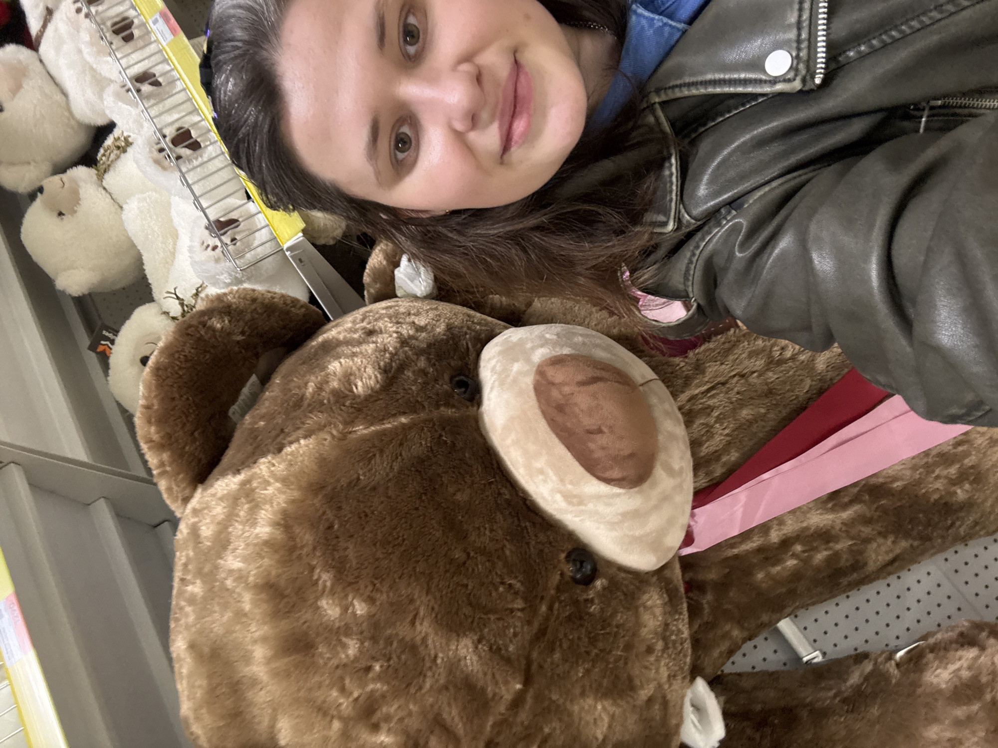 The image displays a female individual, estimated to be between 25-35 years of age, with dark brown hair and brown eyes, captured in a selfie-style photograph. She is positioned on the right side of the frame, looking directly at the camera with a neutral expression and a slight smile. She is wearing a dark, possibly olive-green or grey, leather-style jacket over a blue denim shirt.
To her left, occupying a significant portion of the frame, is a very large brown plush teddy bear. The bear features a lighter brown snout and black button eyes. A pink and a red fabric ribbon or strip are visible around the bear's neck or chest area.
The background indicates an indoor retail environment, specifically a store located in Izhevsk, Russia. White metal shelving units are visible behind the subjects, displaying numerous smaller white plush teddy bears or similar animals. Some of these smaller plush toys appear to have gold-colored accents. Yellow price tag holders are affixed to the front edges of the shelves. The overhead ceiling is white and features a pattern of small, dark perforations, consistent with commercial interior architecture.