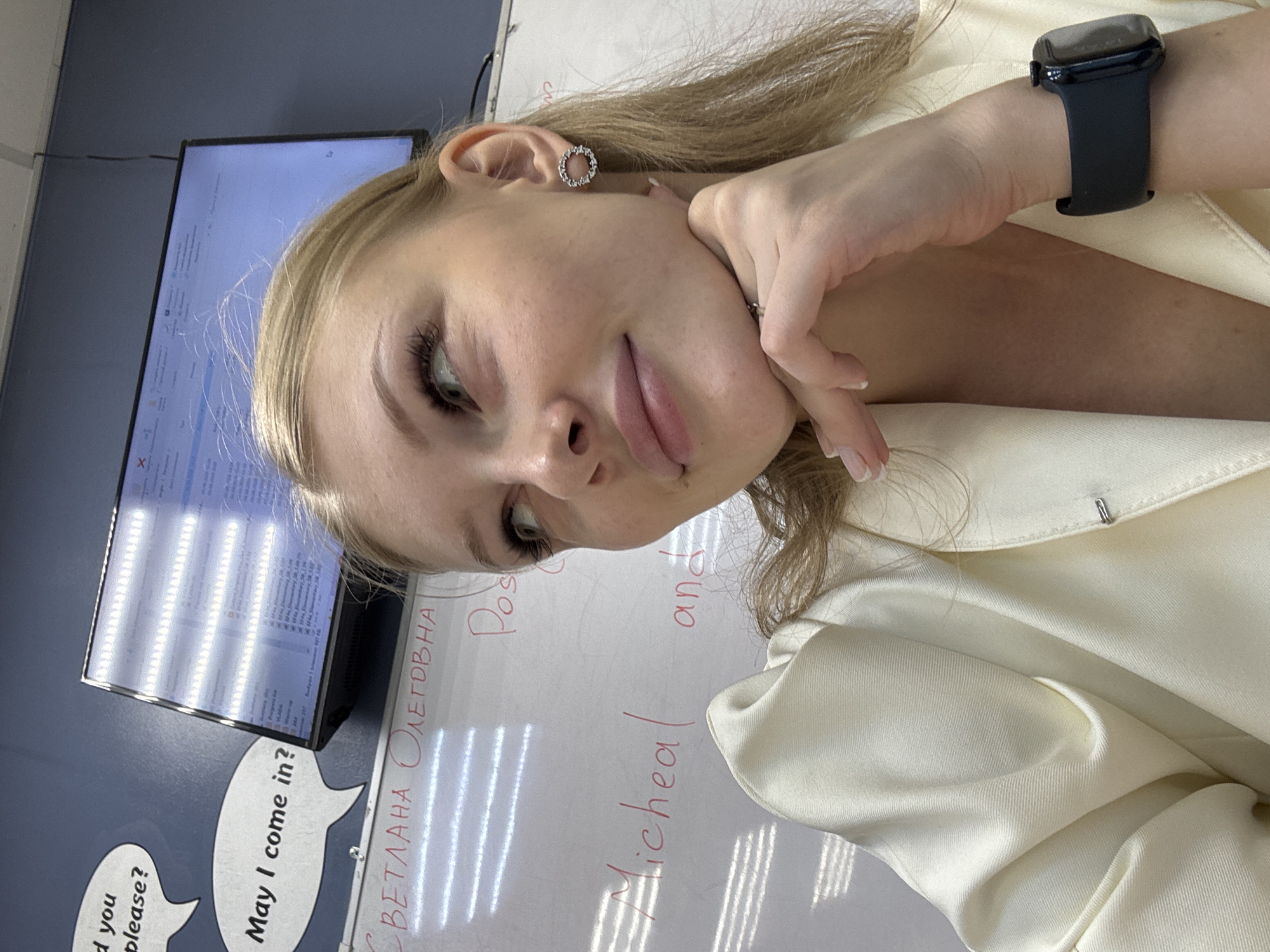 The image is a close-up, waist-up, indoor shot of a young woman with blonde hair and light skin, wearing a light-colored blazer and a black smartwatch. She is looking off to the side, with her chin resting on her hand. Behind her, a whiteboard is visible with writing on it, including Cyrillic text that appears to be a name, "СВЕТЛАНА ОЛЕГОВНА," and the words "Micheal" and "and." There are also some horizontal lines reflecting on the whiteboard, possibly from blinds. In the upper left corner of the image, two speech bubbles are drawn on a blue wall, with the text "d you please?" and "May I come in?". A computer monitor displaying what looks like a file directory is mounted on the wall above the whiteboard. The overall environment appears to be an office or classroom setting, and given the Cyrillic text, it is likely located in Perm, Russia.