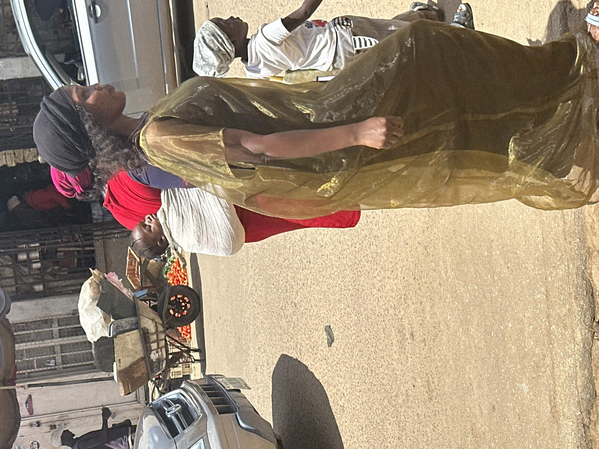 In Narayi, Nigeria, a medium shot captures a street scene with multiple individuals and a vehicle. In the foreground, a woman in a flowing, sheer gold dress walks to the right, her head covered by a black headscarf. To her left, a baby is wrapped in a white cloth and carried on someone's back. Behind them, another person wears a vibrant red garment. Further left, a man in a white cap stands near a cart laden with various items, including what appear to be vegetables. A silver car is parked on the right side of the frame. The ground is paved with a rough, sandy surface, suggesting an outdoor marketplace or busy street. The lighting indicates a bright, sunny day.
