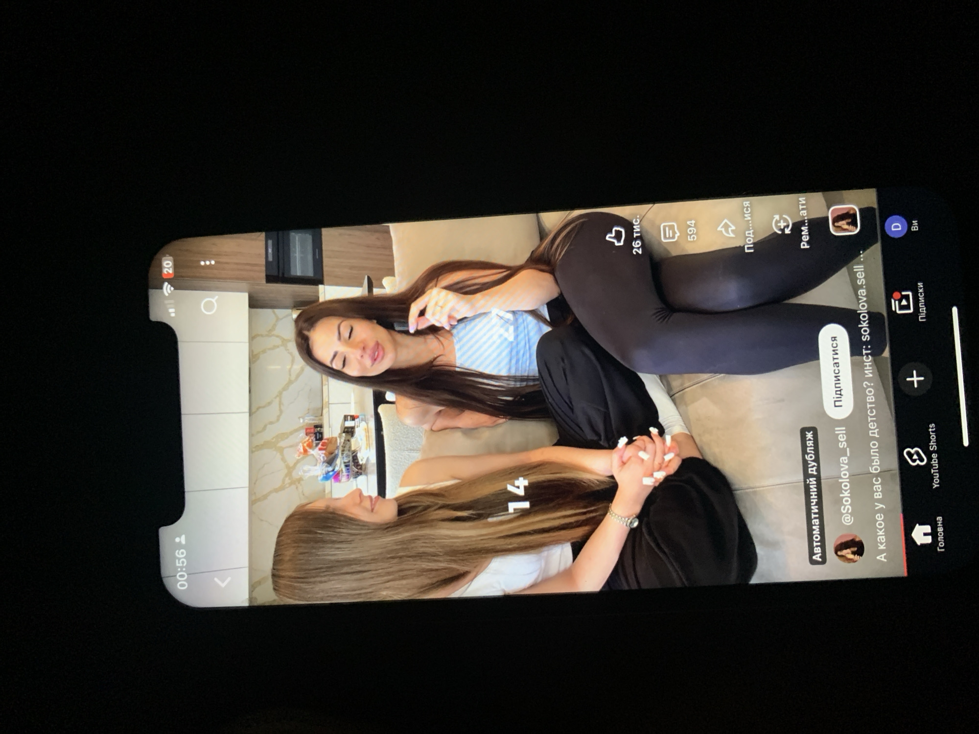 The image shows a close-up view of a smartphone screen displaying a YouTube Shorts video. Two young women are seated on a couch, with one woman on the left and the other on the right. The woman on the left has long, straight, light brown hair and is wearing a white top and black pants. She is looking to her right with her hands clasped in her lap. The woman on the right has long, dark brown hair and is wearing a light blue and white striped top and black leggings. She is holding a small black object in her right hand and looking forward with her eyes closed. The background appears to be a kitchen with white cabinets and marble countertops.
On the smartphone screen, there are several UI elements visible. The top left corner shows "00:56", indicating the video playback time. To the right of the time is a battery icon and a signal strength indicator. In the top right corner, there is a notification icon showing "20". Below the video, there is text that reads "Автоматичний дубляж" (Automatic dubbing) and "@Sokolova_sell Підписатися" (Subscribe). Below this, there is a question in Russian: "А какое у вас было детство?" (What was your childhood like?). On the right side of the video, there are icons for likes ("26 тис." for 26 thousand), comments ("594"), shares, and a refresh icon. The bottom navigation bar of the YouTube app shows icons for "Головна" (Home), "YouTube Shorts", a plus sign, "Підписки" (Subscriptions), and "Ви" (You).
The image is taken from an angle, suggesting it is a photograph of the smartphone screen, possibly captured in a dimly lit environment. There is no visible indication of the location being Zhmerynka, Ukraine, or any other specific city. The content of the video itself does not provide any geographical clues.