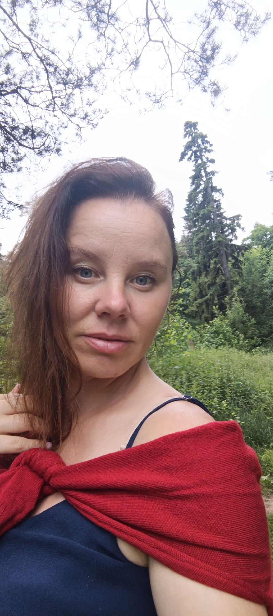 A woman is taking a selfie outdoors in a forest. She is wearing a navy blue top and a red shawl draped over her shoulders. She has long brown hair and blue eyes. The background is a dense forest with tall trees and lush green foliage. The sky is overcast and appears to be a bright white. The image was taken in the city of Chaikovsky, Russia.