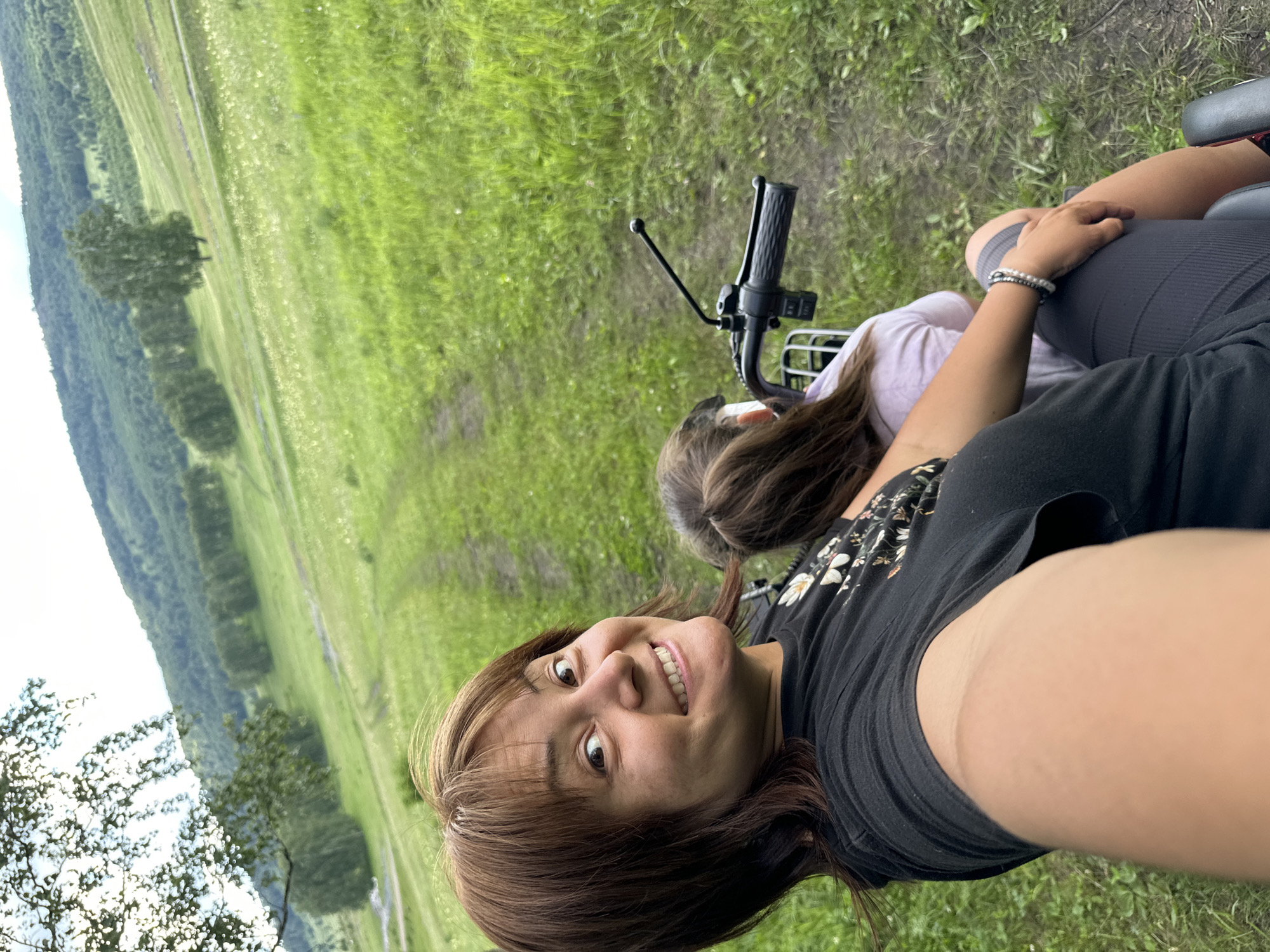 A woman with brown hair and a black t-shirt is riding on an ATV in a grassy area. She is smiling and looking at the camera. A child with brown hair is sitting in front of her on the ATV. The background is a lush green landscape with rolling hills and trees. The woman's left arm is extended towards the camera, holding the camera for a selfie. The child's hair is partially obscuring her face. They are both wearing casual clothing. The ATV's handlebar and controls are visible in the foreground. The overall scene depicts an outdoor recreational activity in a natural setting.