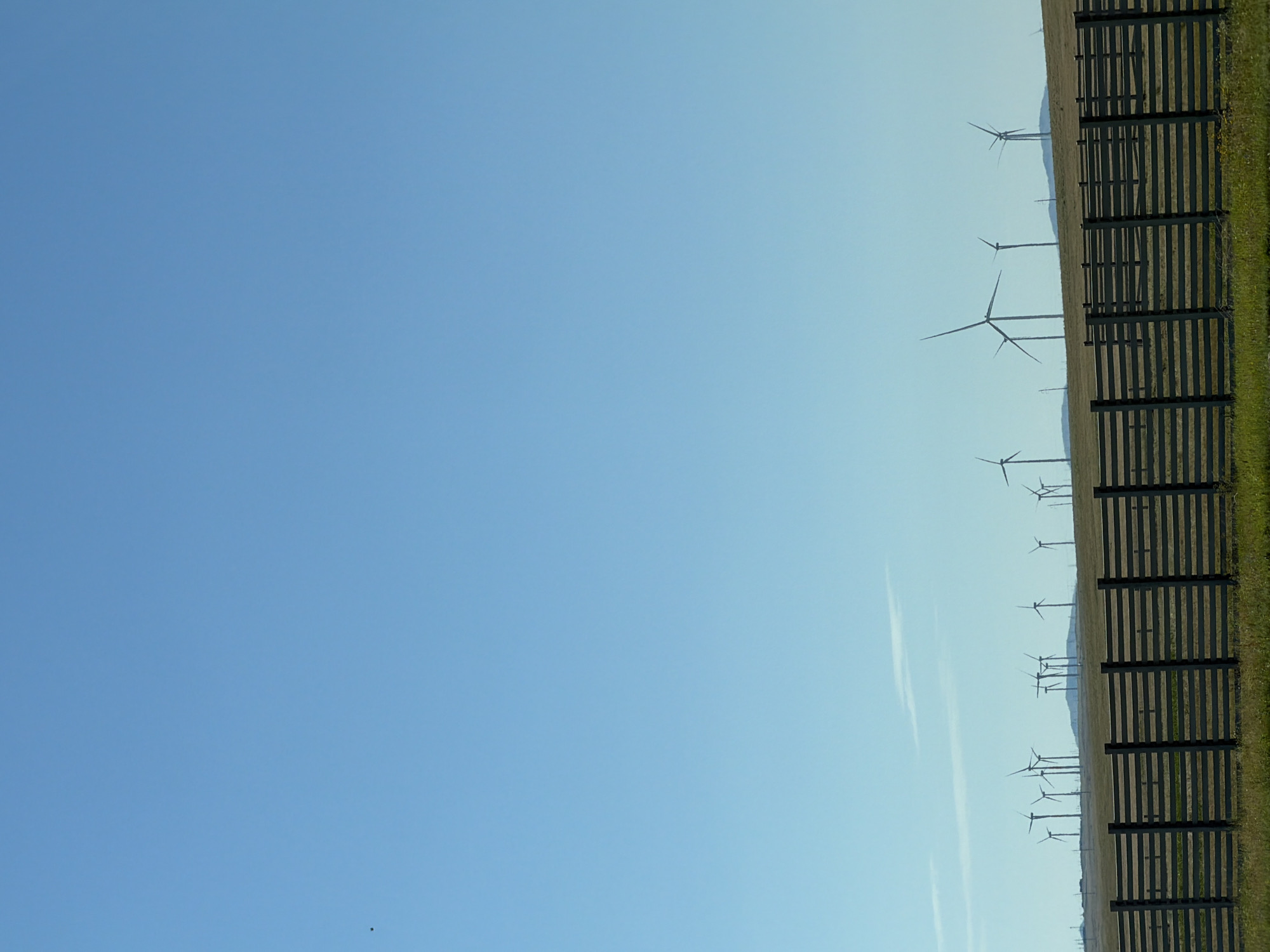 The image depicts a vast, open landscape under a clear blue sky. In the foreground, a weathered wooden fence with horizontal planks stretches across the frame. Behind the fence, a grassy field leads to a distant line of rolling hills. Silhouetted against the sky on the horizon are numerous wind turbines, characteristic of a wind farm. The scene suggests an industrial area integrated into a rural or semi-rural environment. The location is identified as Ereymentaw, Kazakhstan. There are no people visible in the image, and the wind turbines are stationary, indicating no current activity related to them. The environment is arid and expansive.