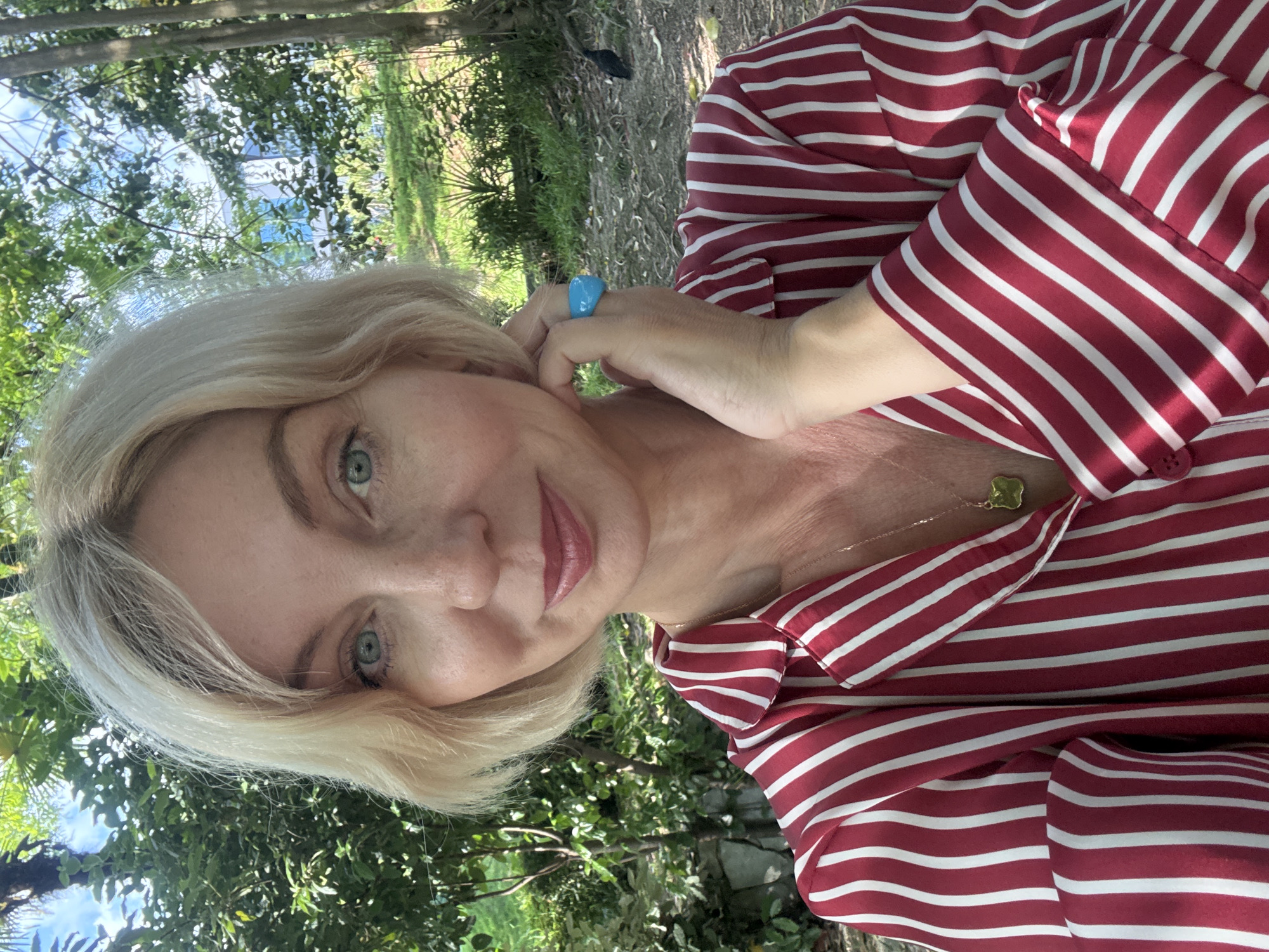 The image shows a selfie of a blonde woman with blue eyes, wearing a red and white striped shirt, a gold necklace, and a turquoise ring on her right index finger. She is outdoors, with lush green foliage and trees in the background, suggesting a natural or park-like setting. The lighting indicates it is daytime, with the sun casting shadows. The environment appears to be a garden or a wooded area, with hints of a building or structures in the distant background. There is no specific indication of the location being Sochi, Russia.