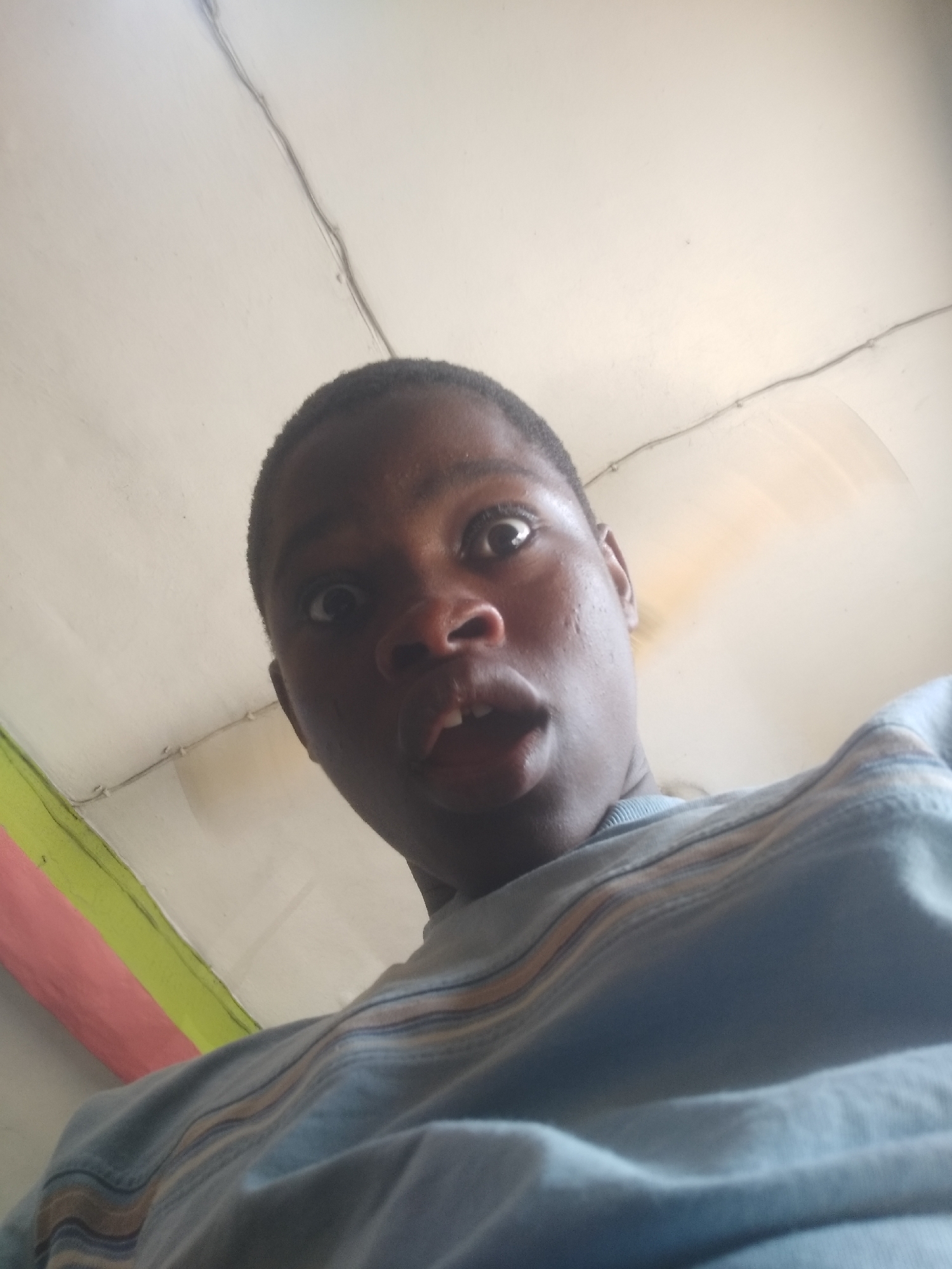 A person with dark skin and short hair is looking upwards with a surprised expression. They are wearing a light blue striped shirt. The background is a white ceiling with exposed wiring and a painted beam in pink, green, and yellow. The location is Shangisha, Nigeria.