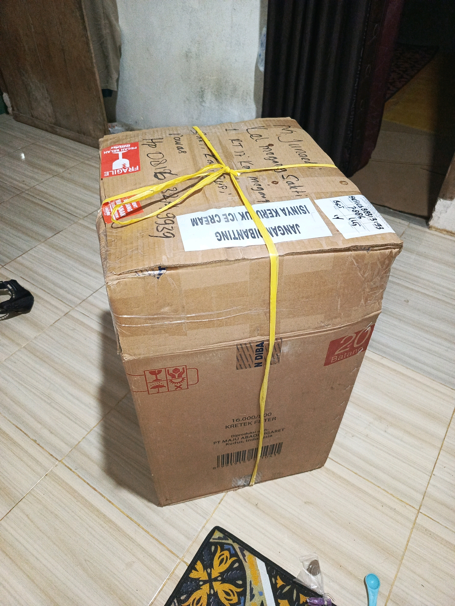 The image shows a cardboard box on a tiled floor. The box is brown and has yellow tape securing it. There are several labels on the box with writing in Indonesian. One label says "FRAGILE" in red and white. Another label has writing that appears to be "JANGAN DIBANTING KERUPUK ICE CREAM" which translates to "DO NOT THROW ICE CREAM CRACKERS". The box also has a label that says "20 BATANG" which translates to "20 STICKS". The background shows a wooden surface and a dark curtain. A patterned mat is visible on the floor in the lower part of the image. Based on the label "DIPRODUKSI DI: PT MAJU ABADI INDONESIA KUDUS, INDONESIA" it is possible this item was manufactured in Kudus, Indonesia, but it is not possible to determine the specific location within Megang Sakti I from the provided image.